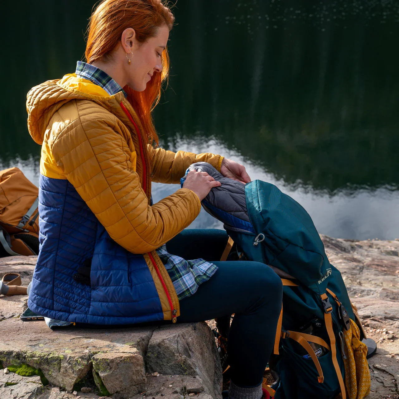 Woman in yellow and blue puffer jacket packing hiking backpack by a lake