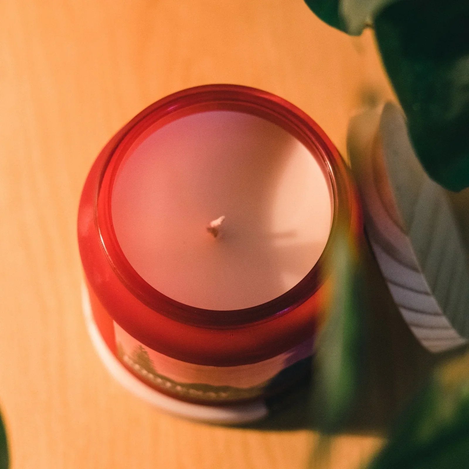 Red glass jar candle on wood table, surrounded by green leaves, top view