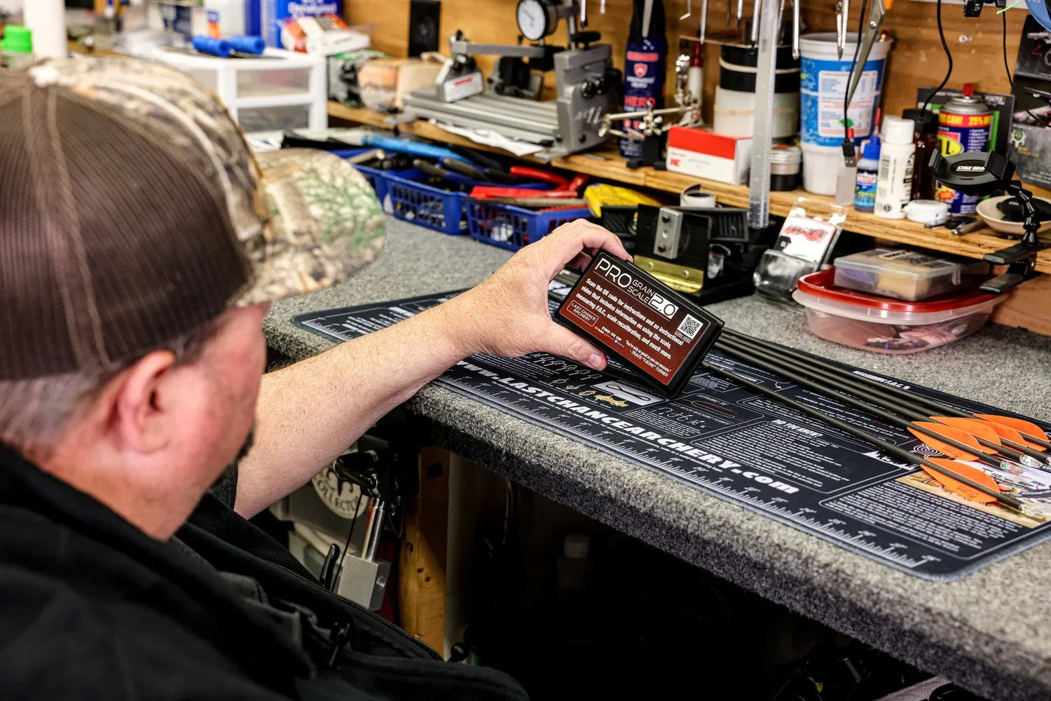 Man in camo hat holding Pro Grain Scale 2.0 at archery workbench with arrows and tools