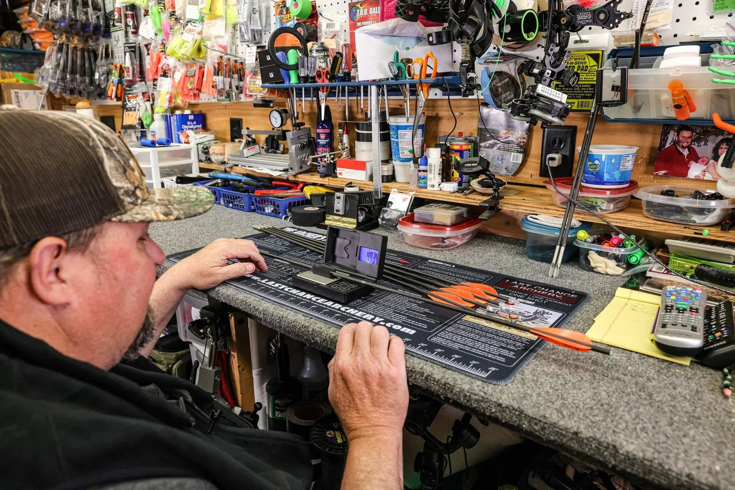 Man at workbench weighing arrows, archery tools and supplies visible on organized shop shelves.