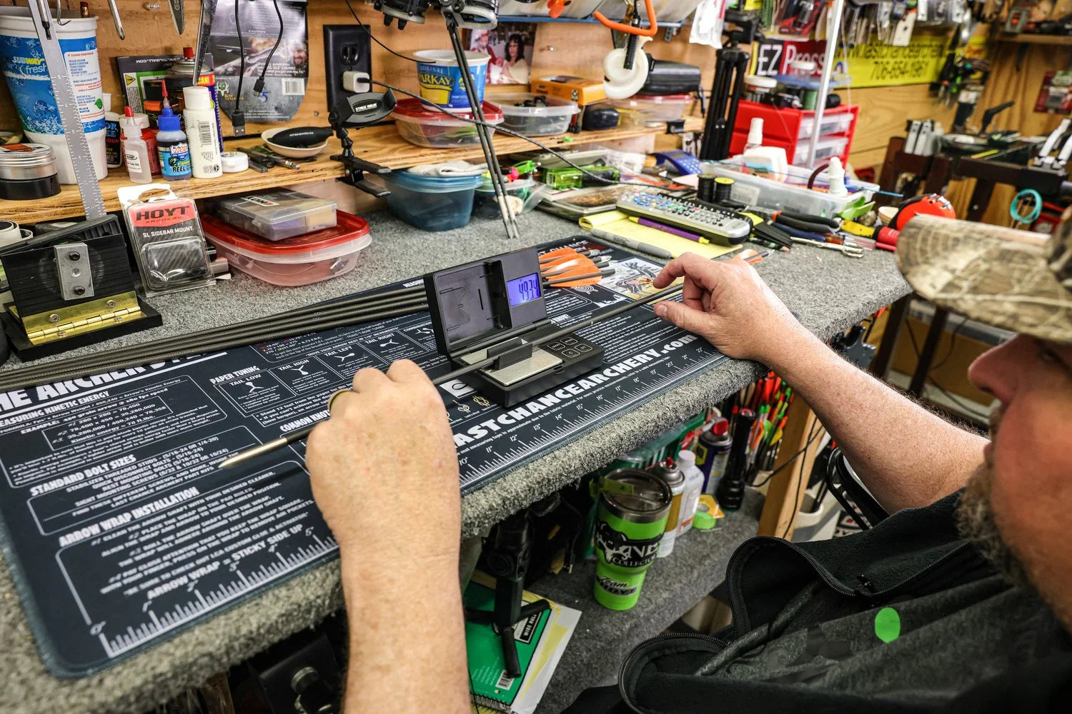 Man measuring arrow weight on digital scale at archery workbench with tools and supplies