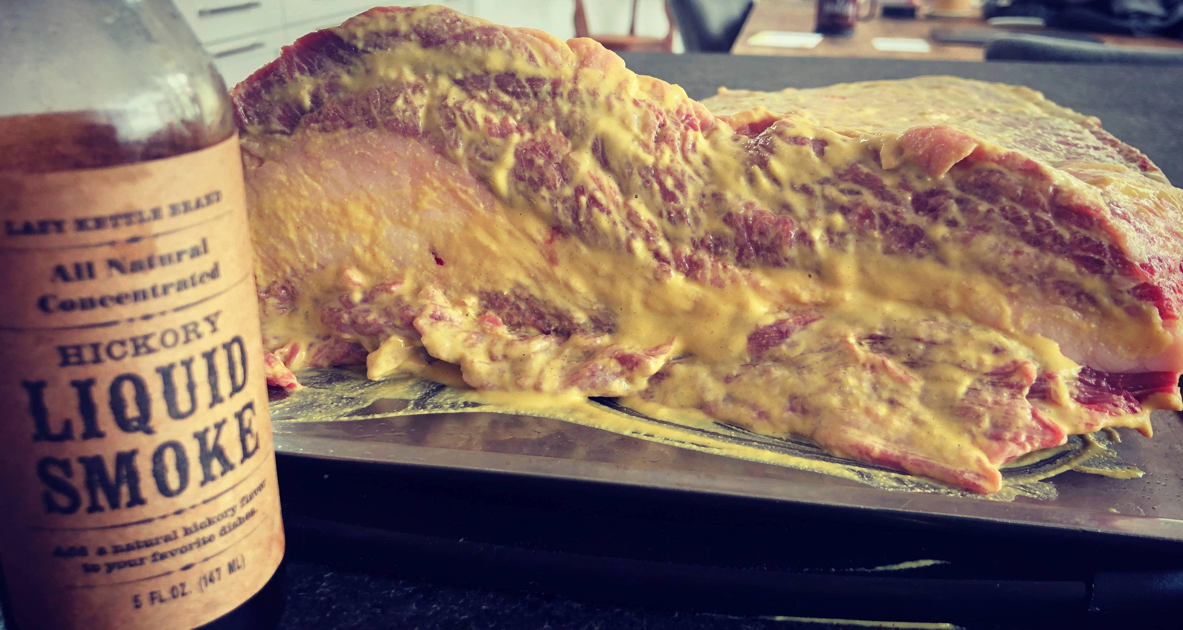 Lazy Kettle Brand Hickory Liquid Smoke bottle next to raw mustard-coated brisket on tray
