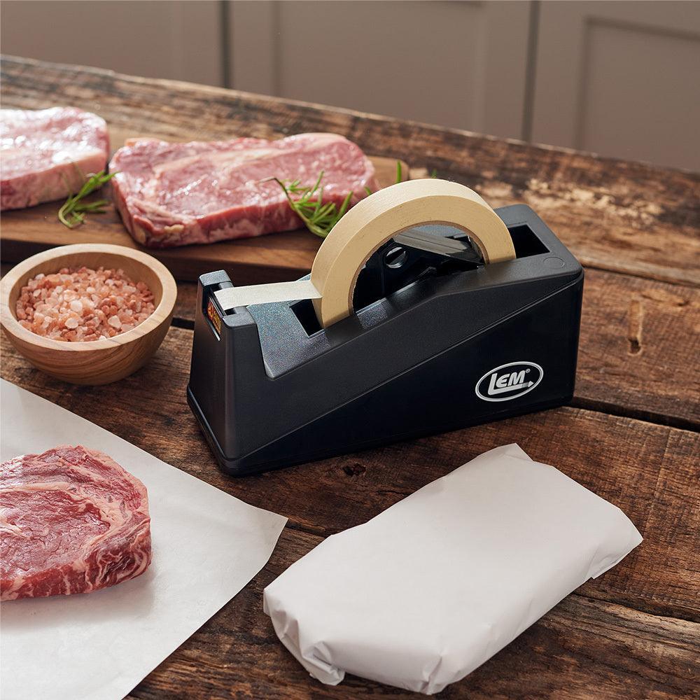 LEM tape dispenser with roll on rustic wood table near steaks, butcher paper, and pink salt bowl