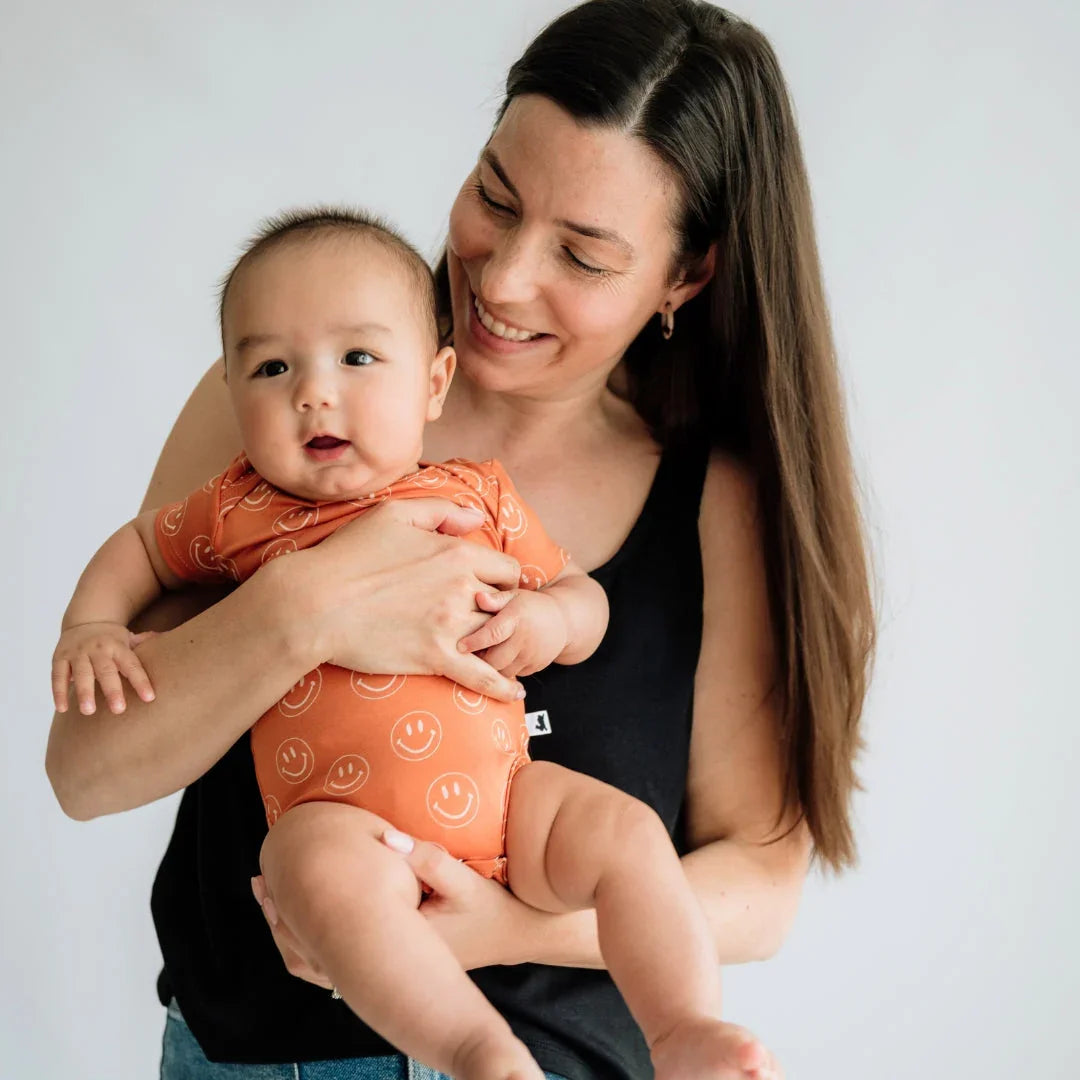 Smiling woman holding baby in orange smiley face onesie against plain background