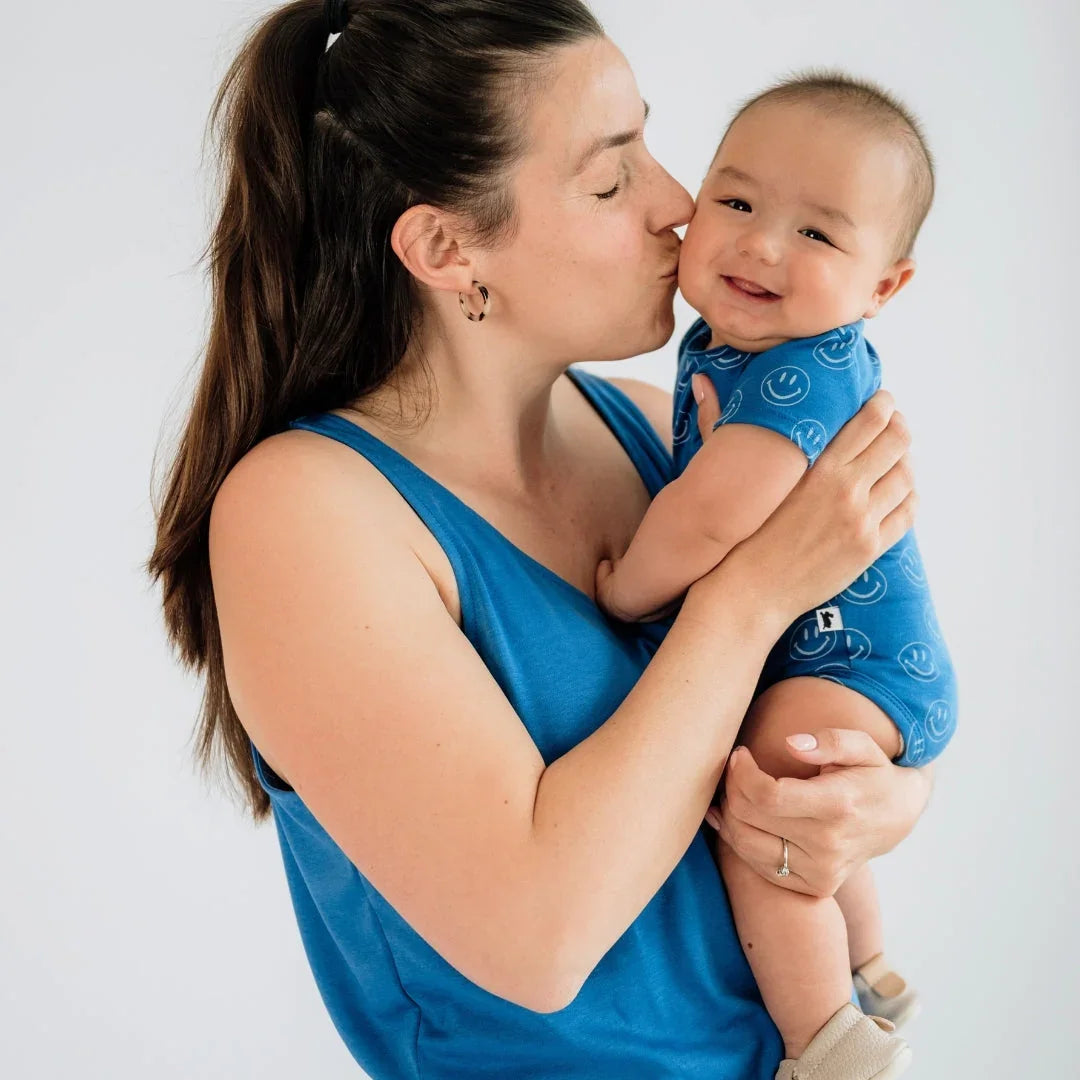 Smiling mother in blue tank top kisses baby in blue onesie with smiley faces on white background