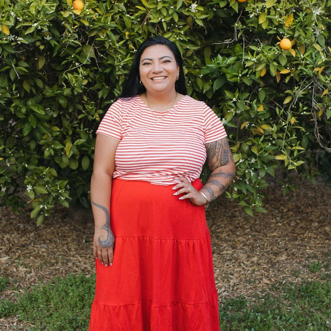 Smiling woman in red striped shirt and skirt stands in garden with fruit tree background