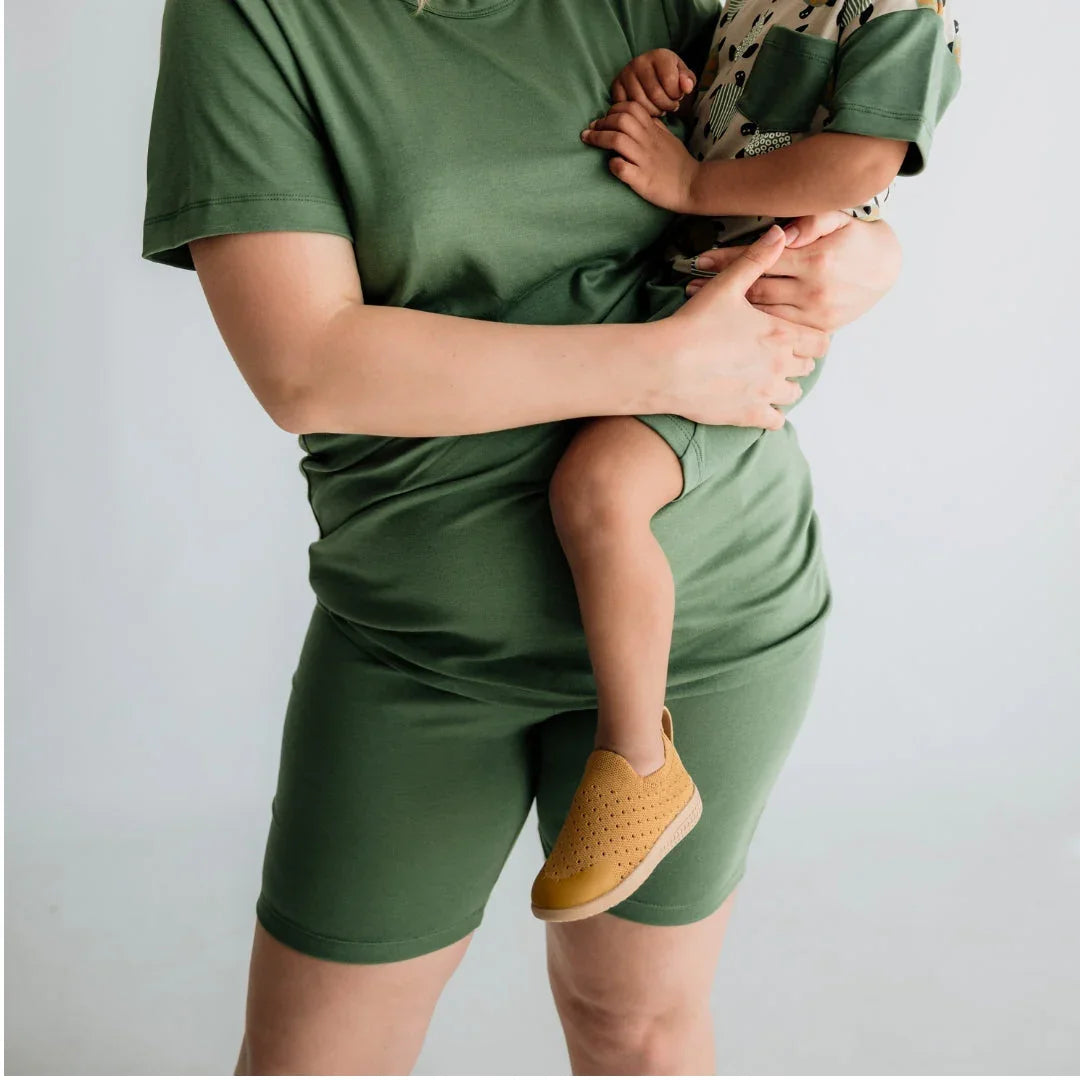Woman in green shorts and t-shirt holding child in printed shirt and yellow slip-on shoe