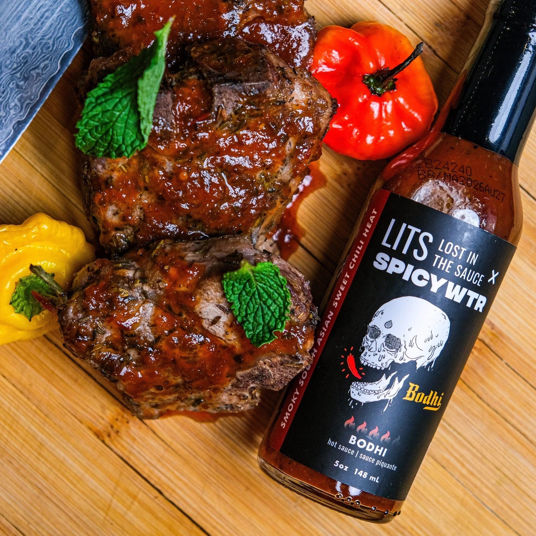 Grilled lamb with chili sauce, mint leaves, hot peppers, and LITS Bodhi spicy hot sauce on wood board