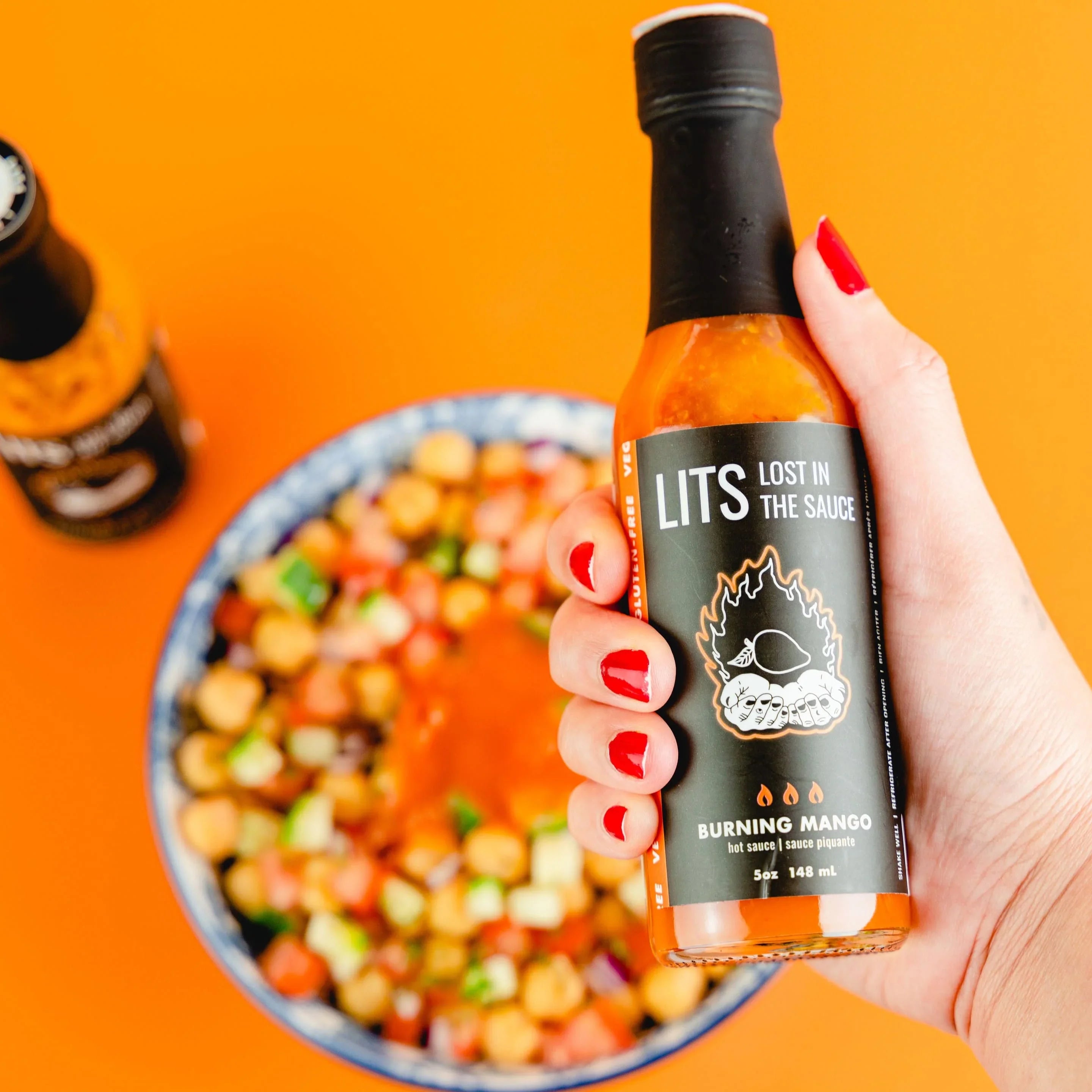 Hand holding LITS Burning Mango hot sauce over a chickpea salad on orange background