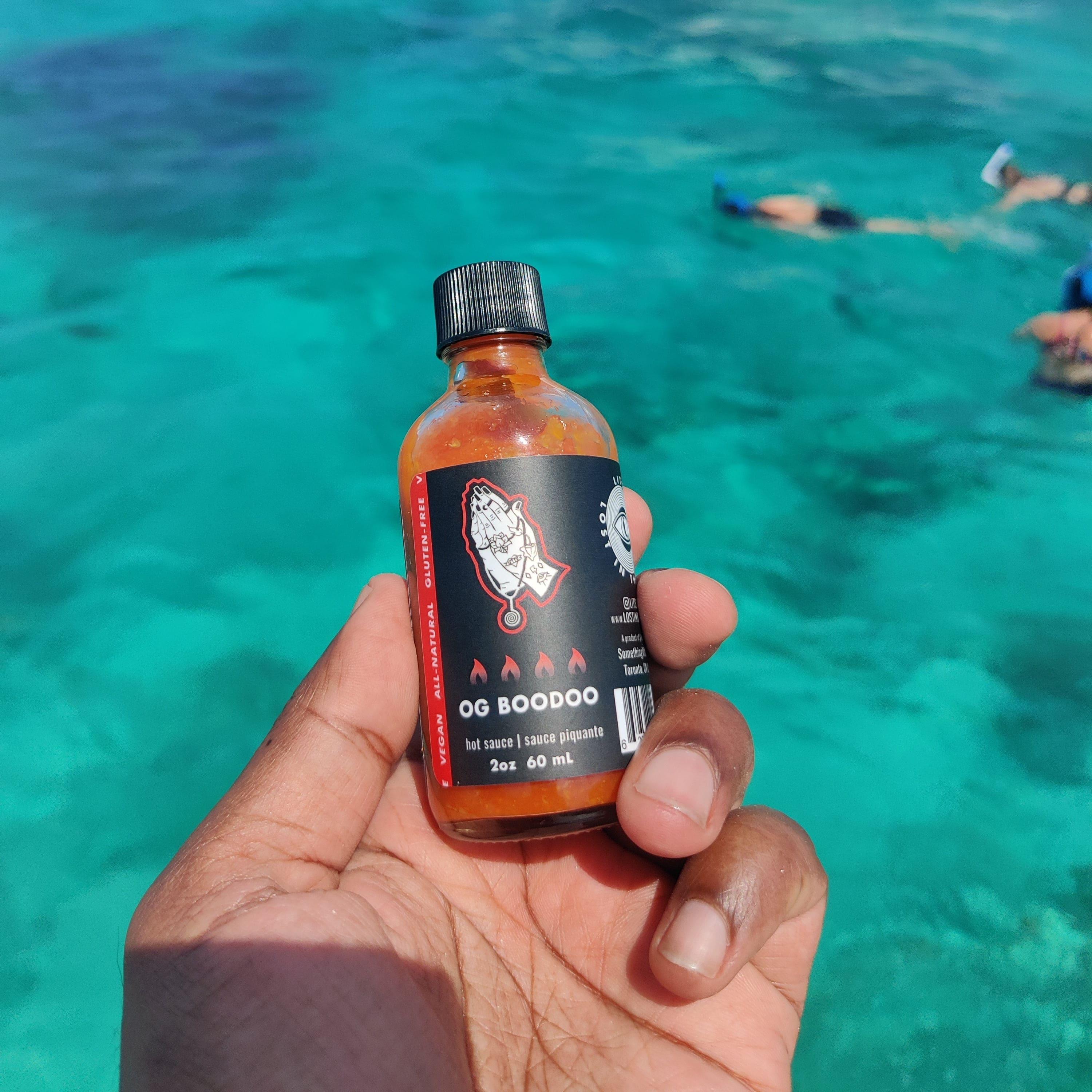 Hand holding OG Boodoo hot sauce bottle above clear turquoise water with snorkelers
