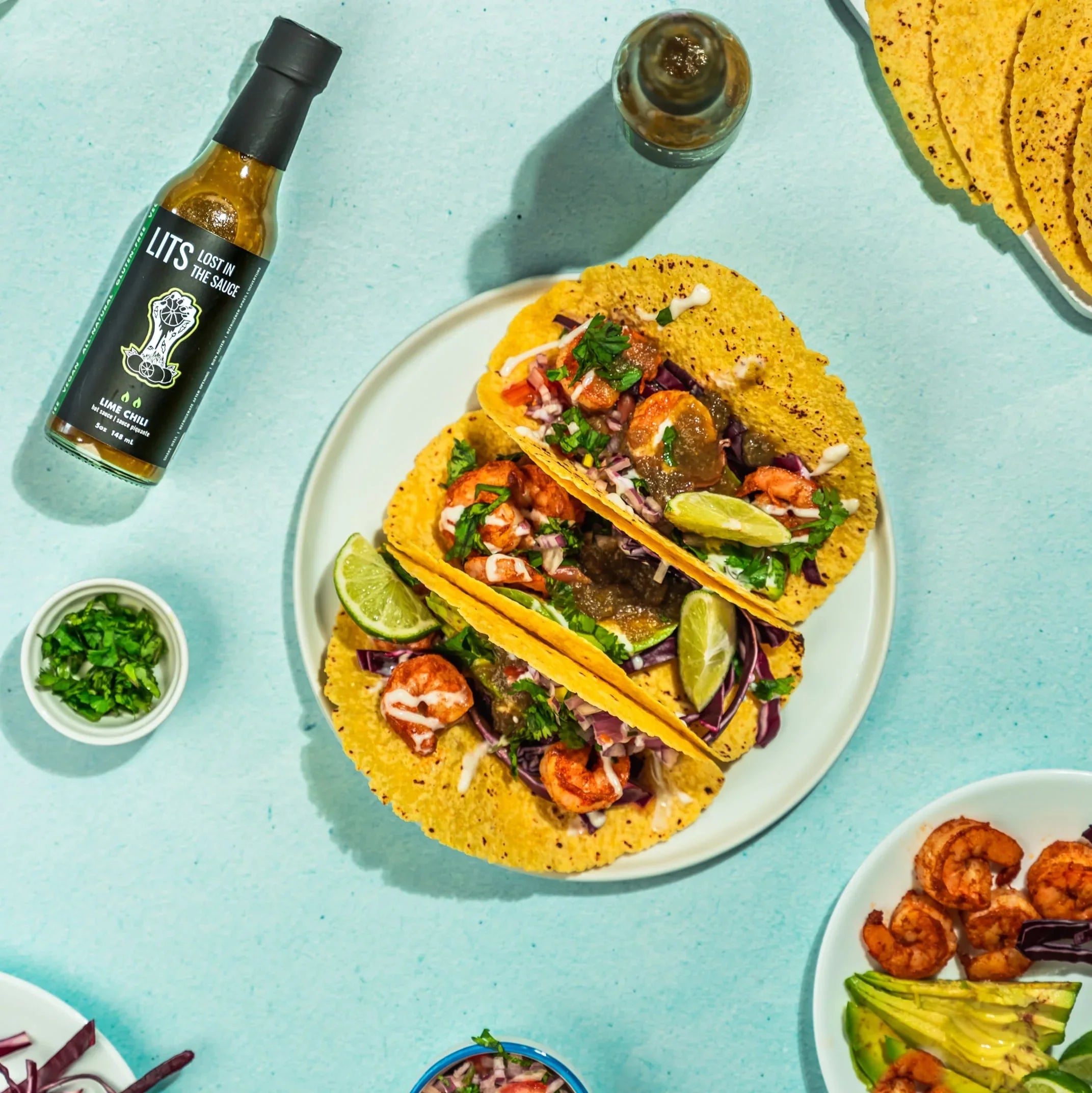 Lime chili shrimp tacos with sauce bottle, tortillas, lime, and cilantro on blue background