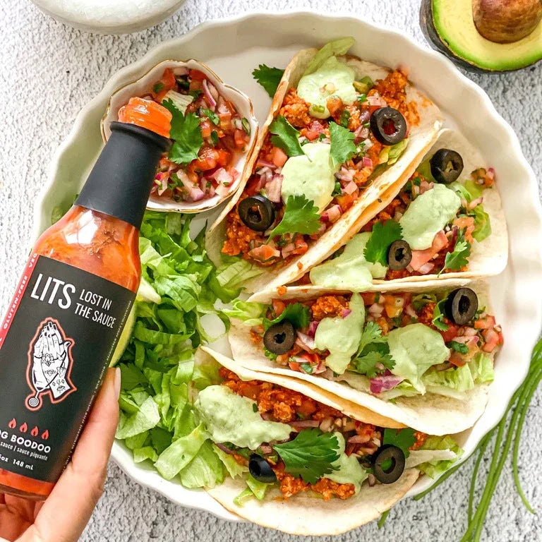 Three tacos with lettuce, salsa, olives, green sauce and a hand holding LITS OG BooDoo hot sauce