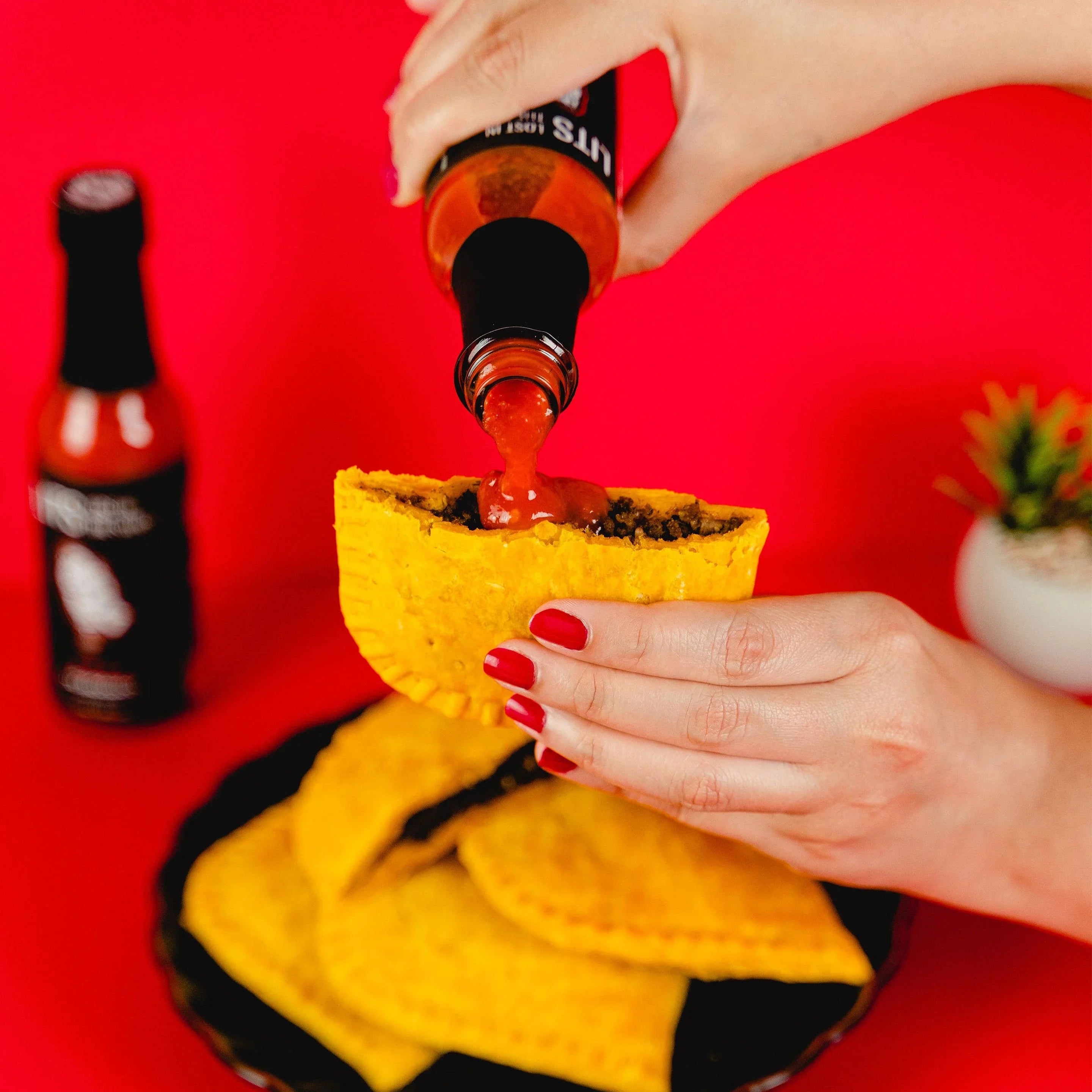 Hot sauce poured on Jamaican patty with plate of patties, red background, woman's hands