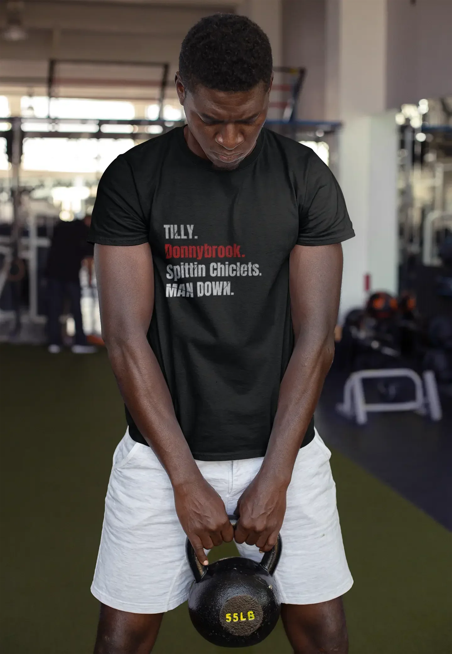 Man in gym holding 55lb kettlebell, wearing black graphic T-shirt and white shorts