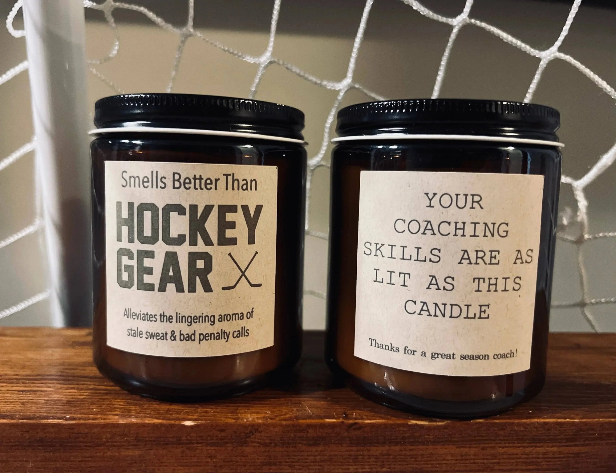 Two brown candles with humorous hockey-themed labels on a wooden shelf in front of a net.