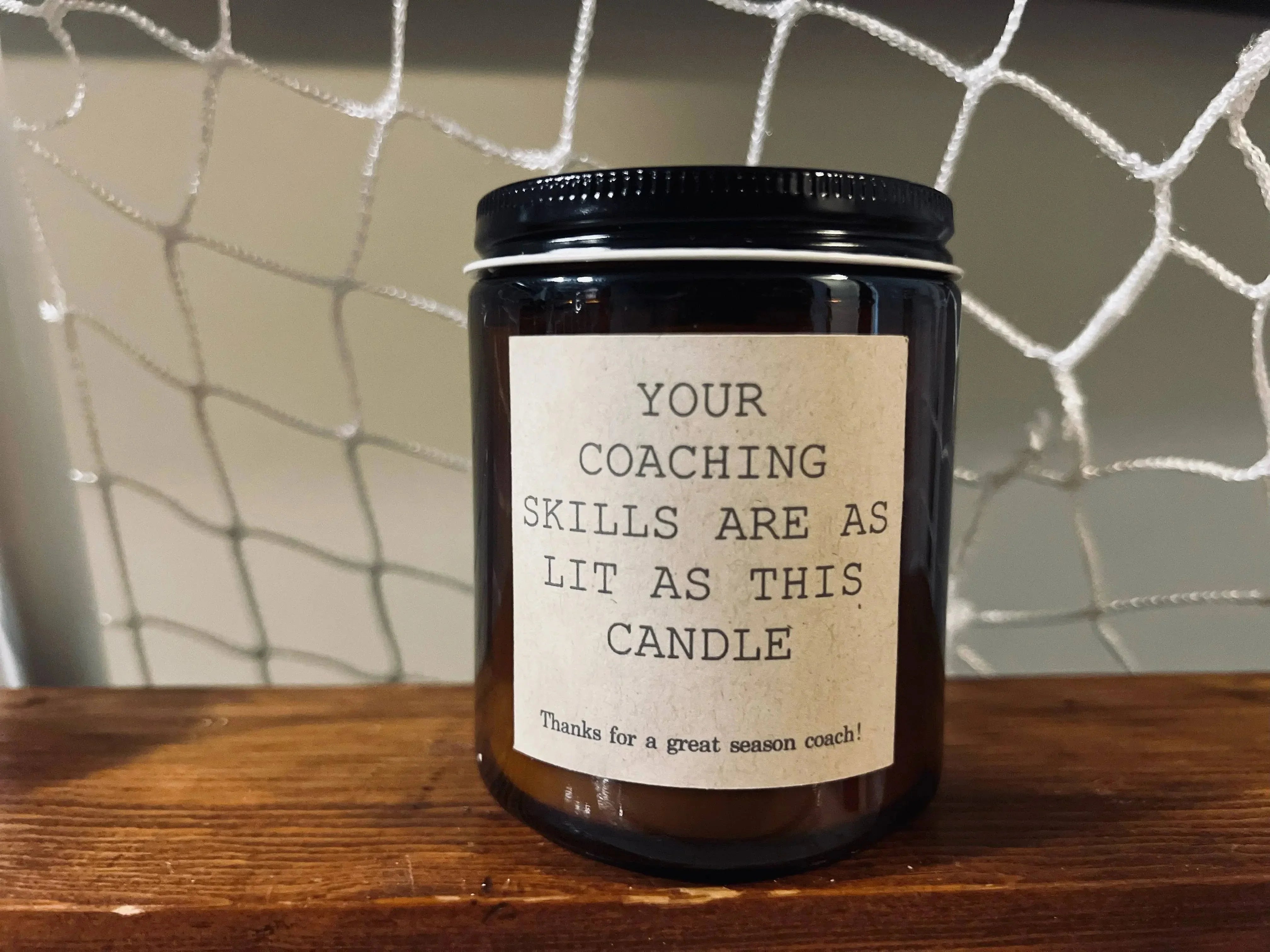 Brown glass candle jar with 'Your coaching skills are as lit as this candle' label, on wood shelf