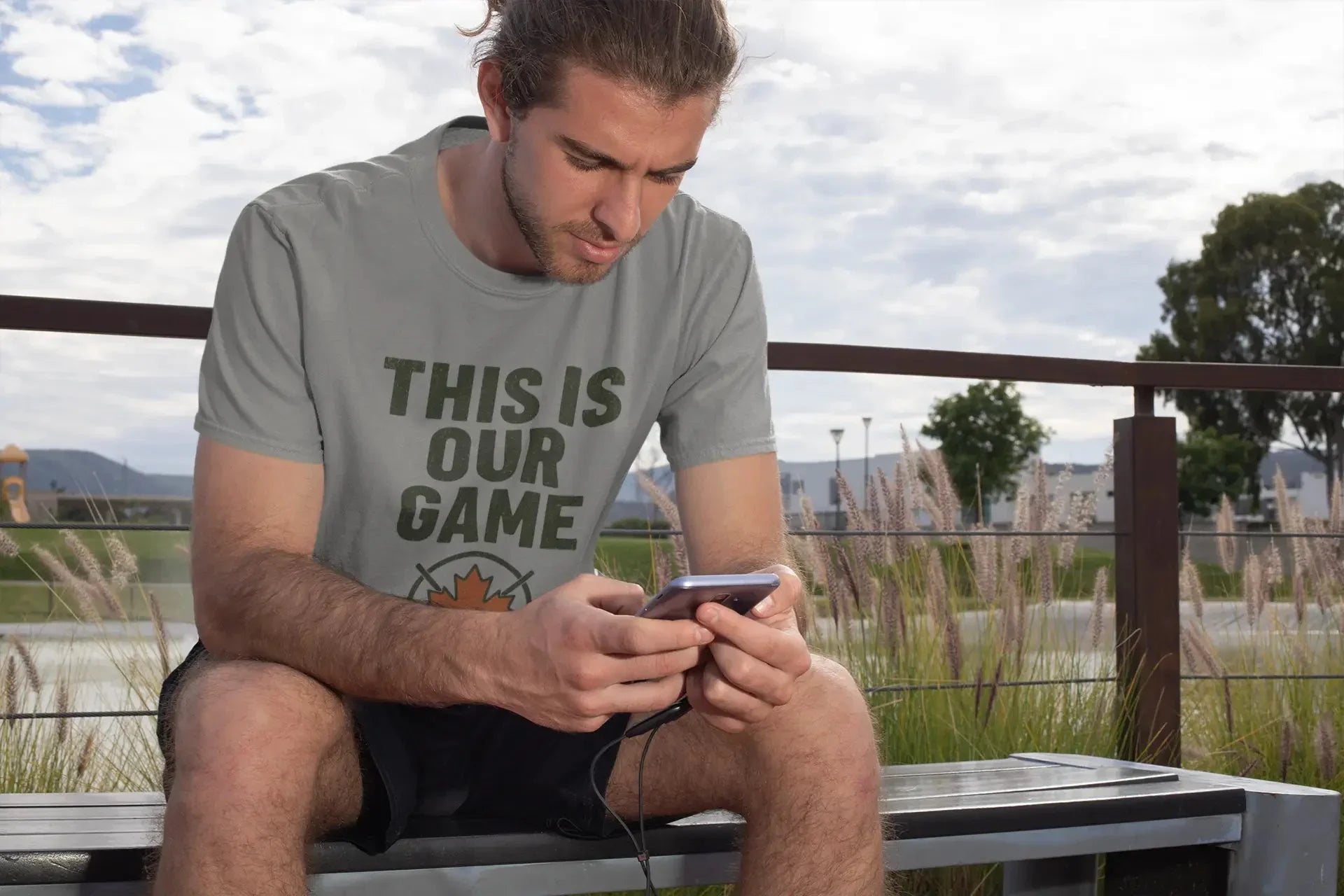 Man in grey 'This Is Our Game' t-shirt using smartphone on outdoor bench