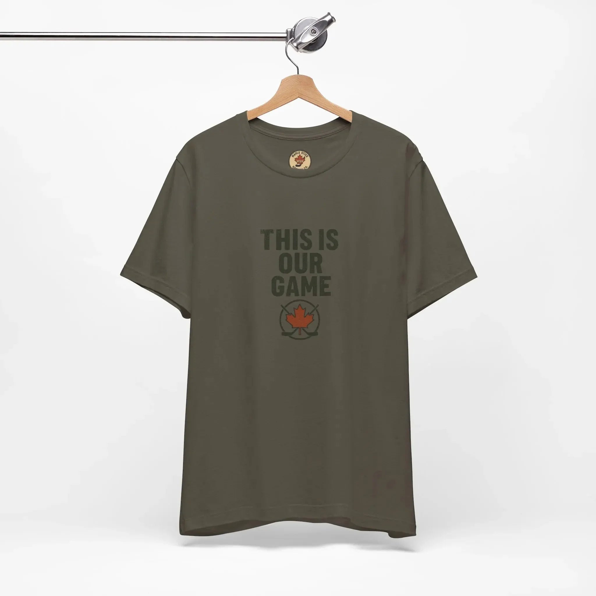 Olive green t-shirt with 'This Is Our Game' text and red maple leaf logo, hanging on wooden hanger
