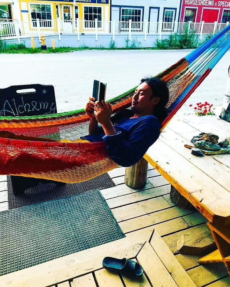 Person relaxing on a colorful Mayan XL hammock reading outside a shop with wooden deck