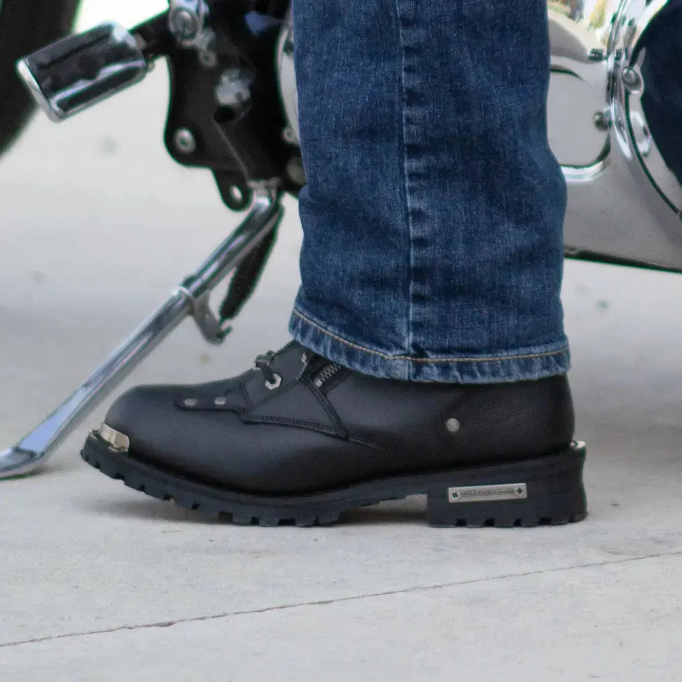Black motorcycle boot with metal toe cap, blue jeans, near chrome motorcycle parts
