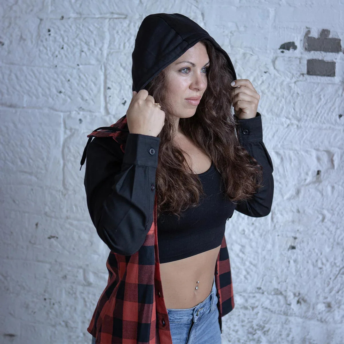 Woman in a red plaid hooded shirt, black crop top, and jeans posing by a white brick wall