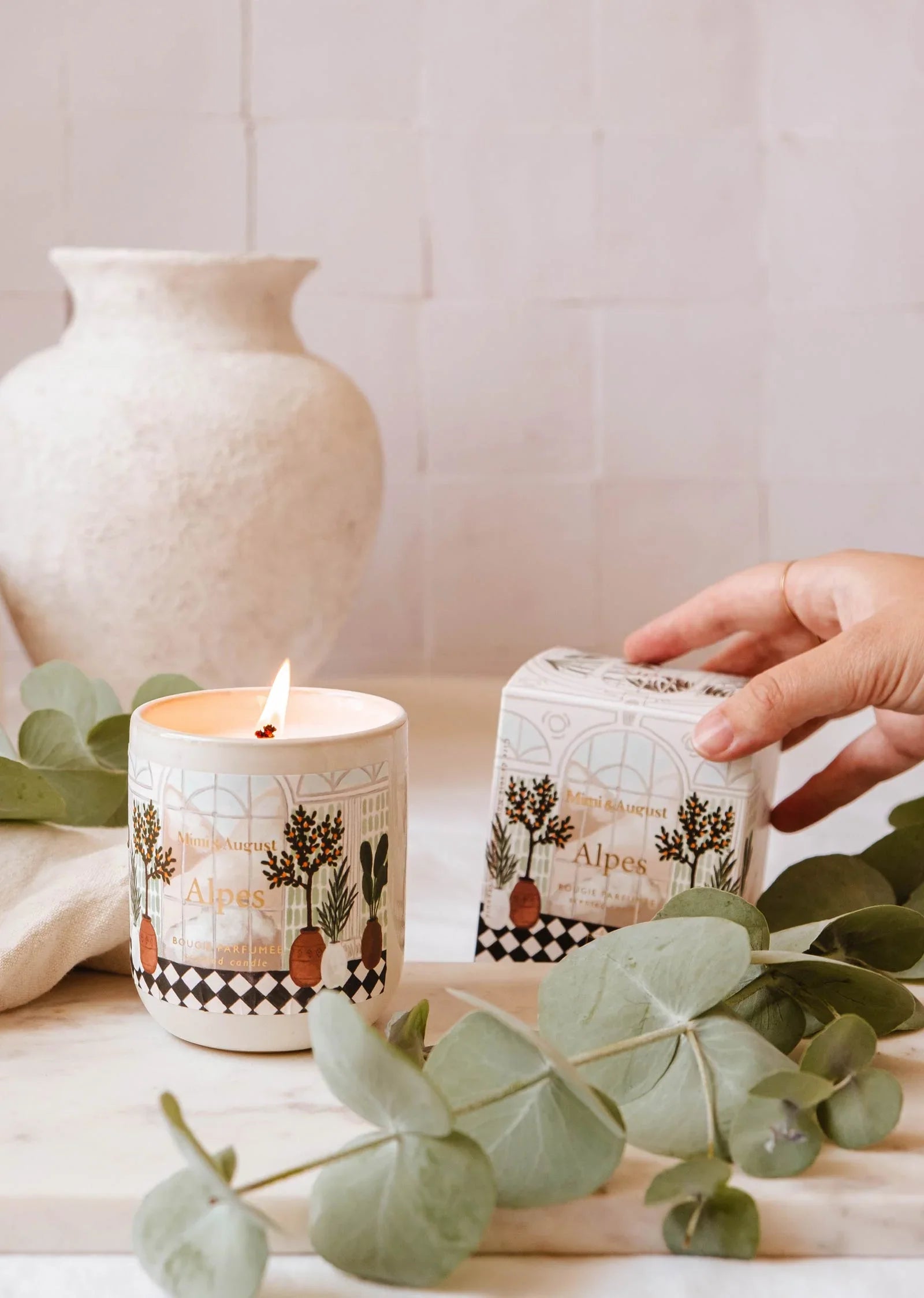Alpes reusable candle by Mimi & August, lit on a marble surface with greenery and box