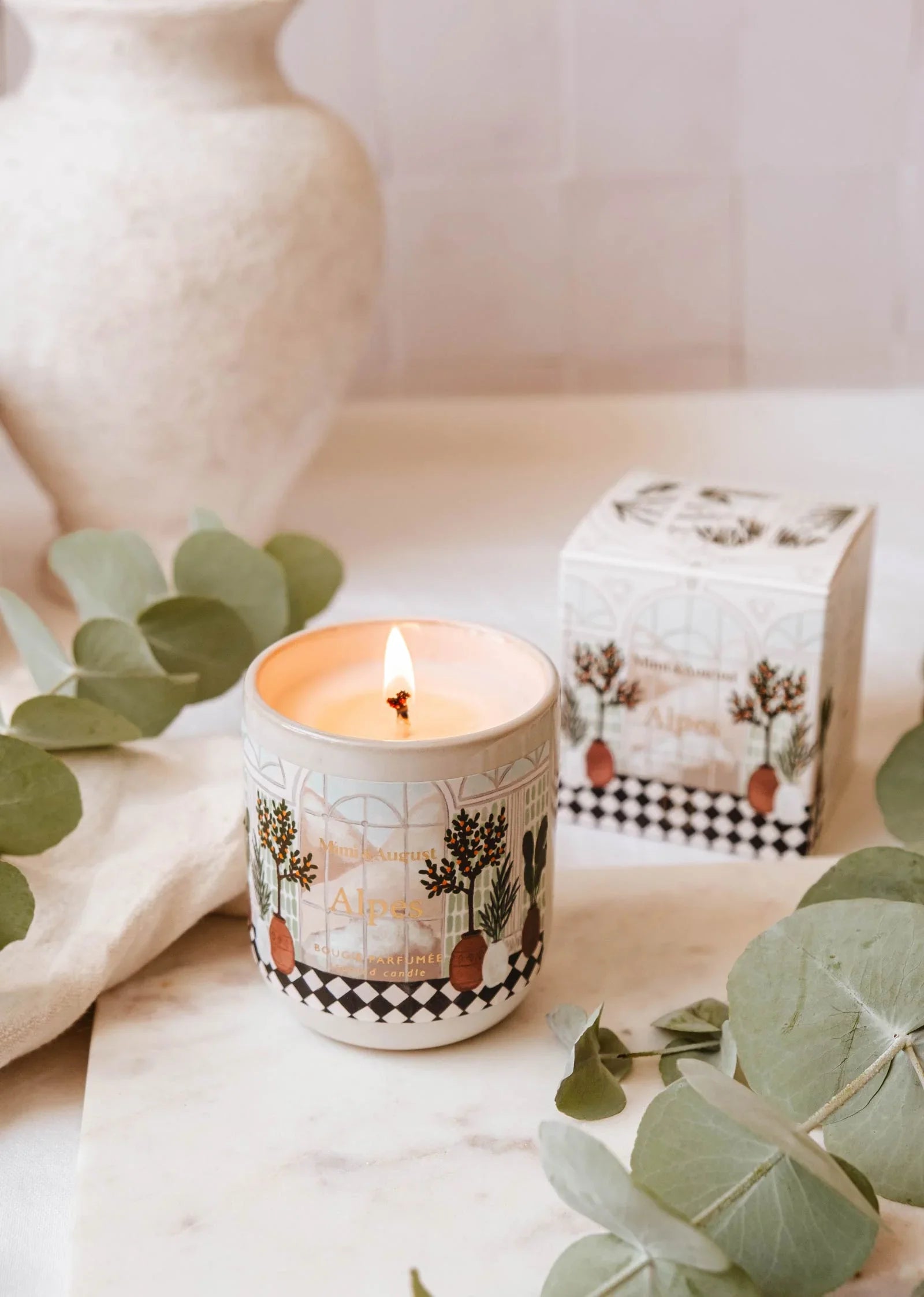 Alpes reusable 8oz candle by Mimi & August, lit, with illustrated ceramic jar and box on a marble surface, surrounded by eucalyptus leaves.