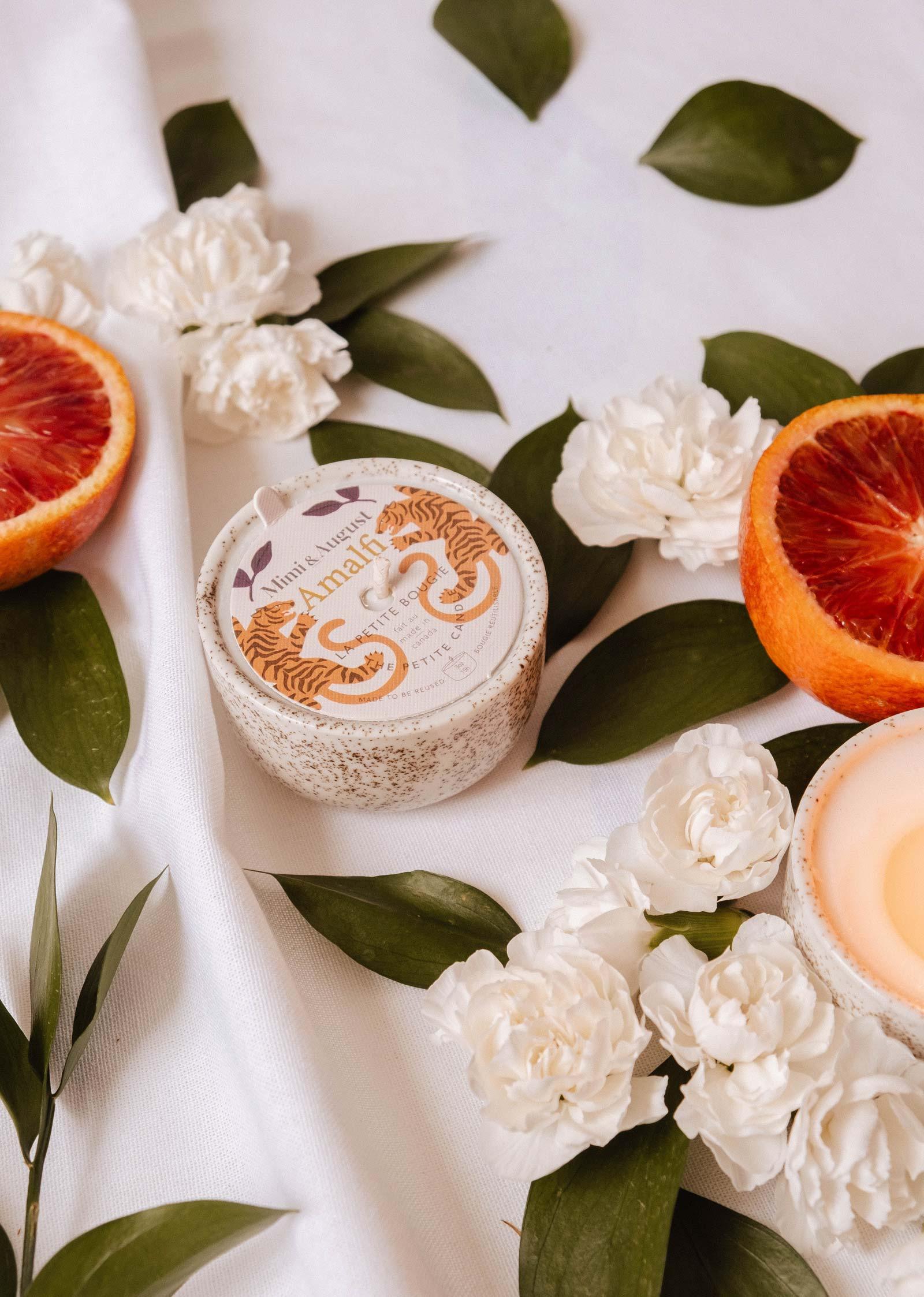 Amalfi reusable candle in a speckled jar, surrounded by white flowers, green leaves, and blood orange halves on white fabric.