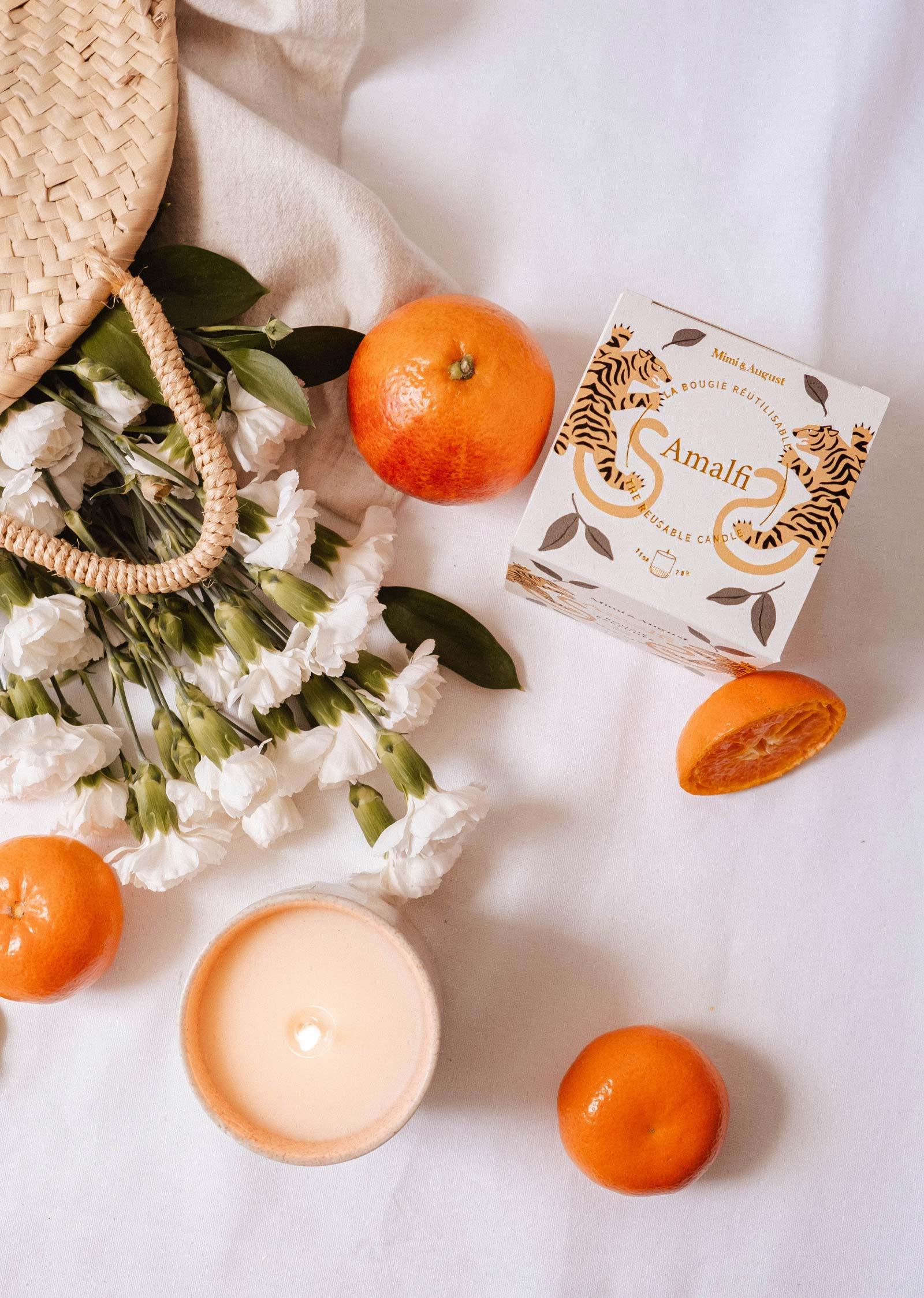 Amalfi reusable candle with oranges, cut orange, white flowers, and a woven bag on cloth