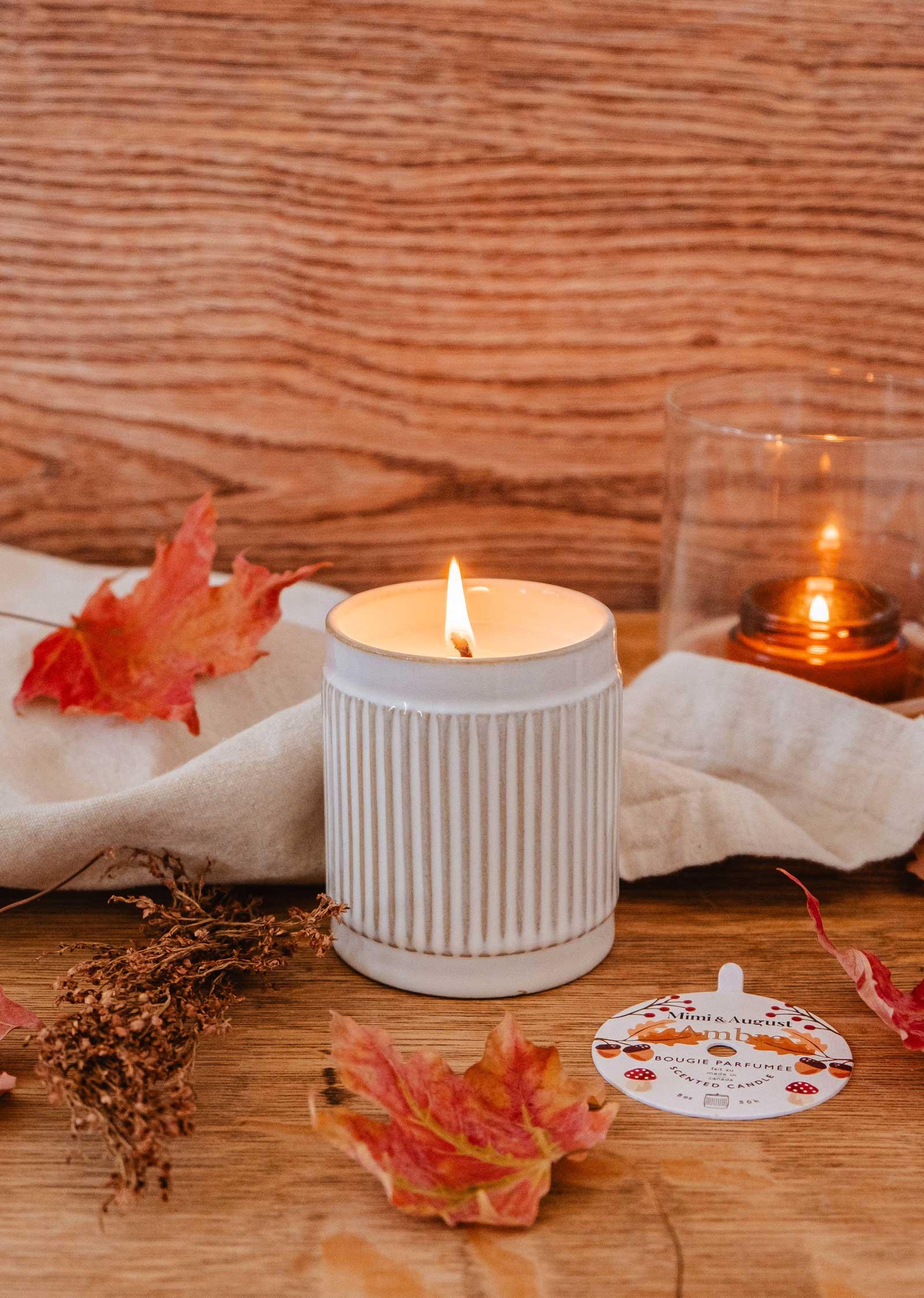 Lit Ambre reusable candle in ribbed white jar on wooden table with autumn leaves and label