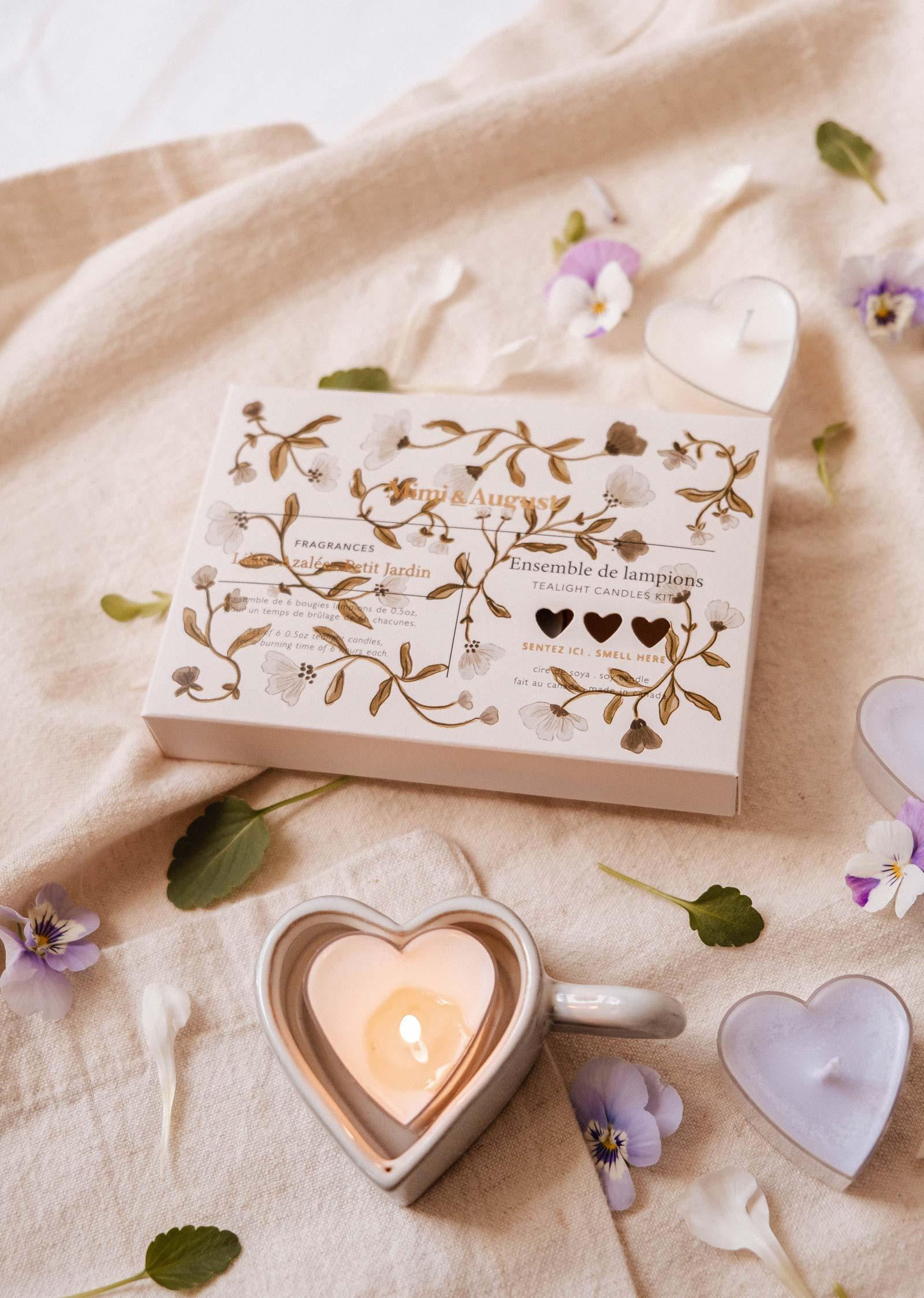 Blush heart-shaped tealight candle set with floral packaging on beige fabric, surrounded by flowers