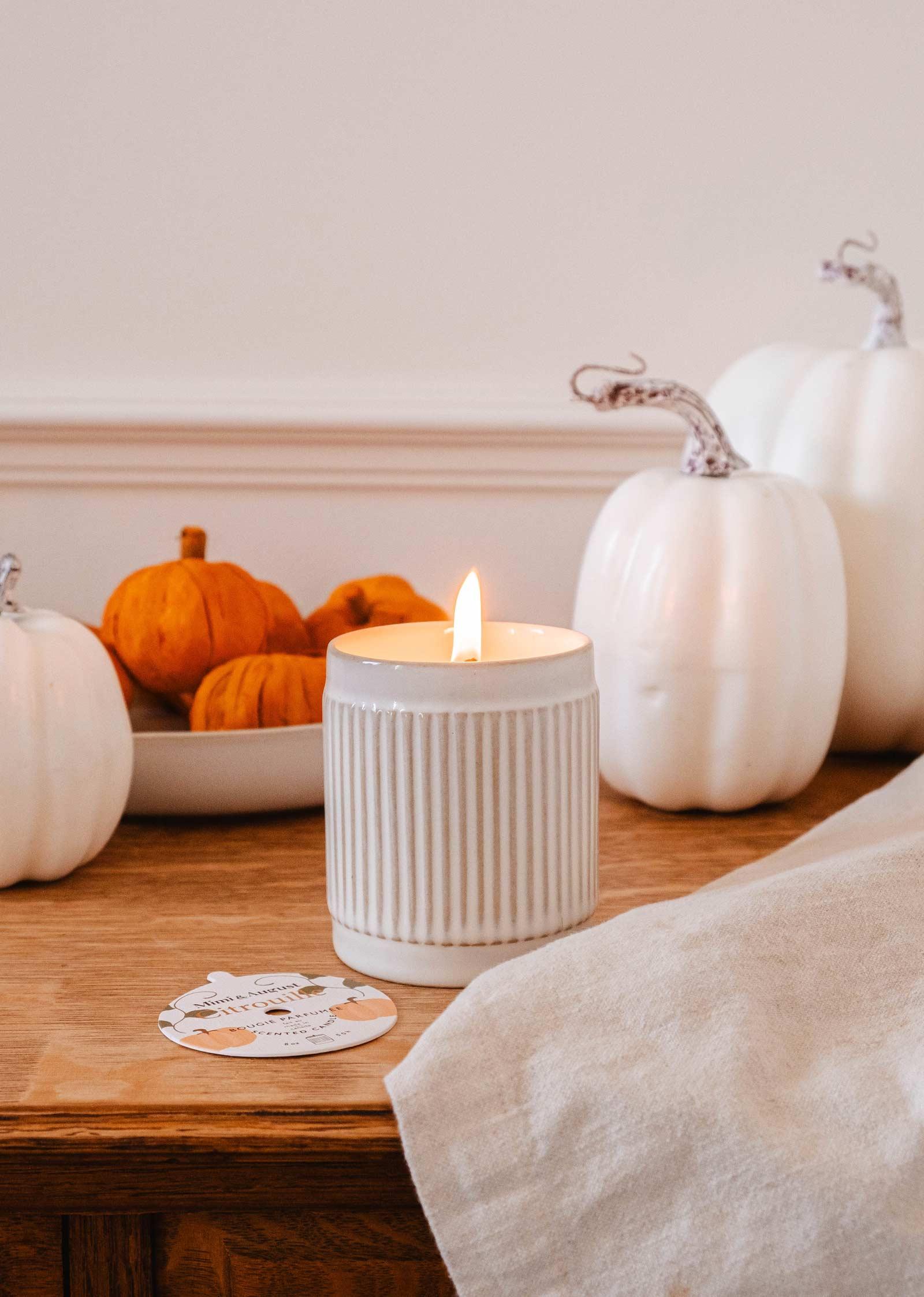 Lit ribbed ceramic candle on wooden table with white and orange pumpkins in a cozy autumn setting