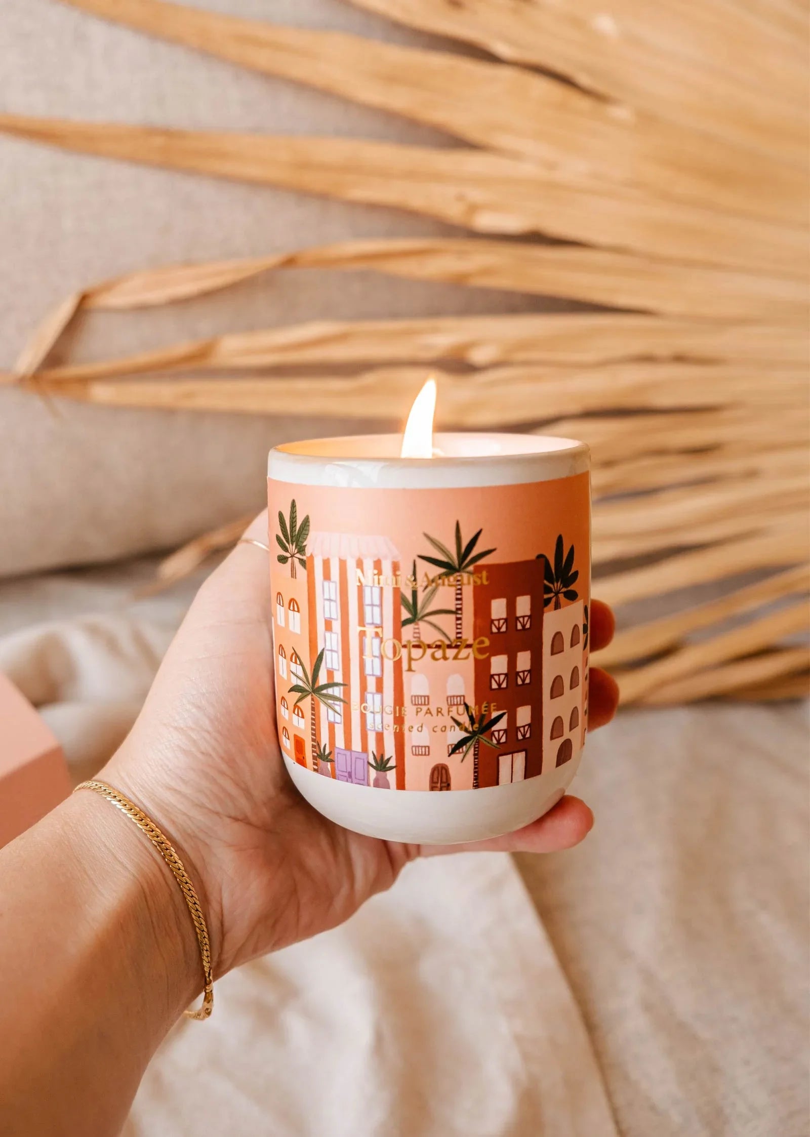 Hand holding Topaze reusable scented candle with orange cityscape design, lit flame