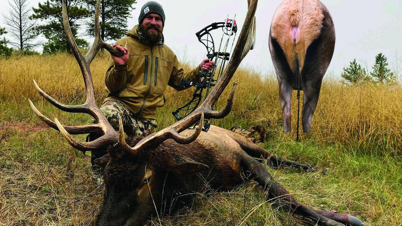Hunter with bow posing next to large elk in grassy backcountry wilderness