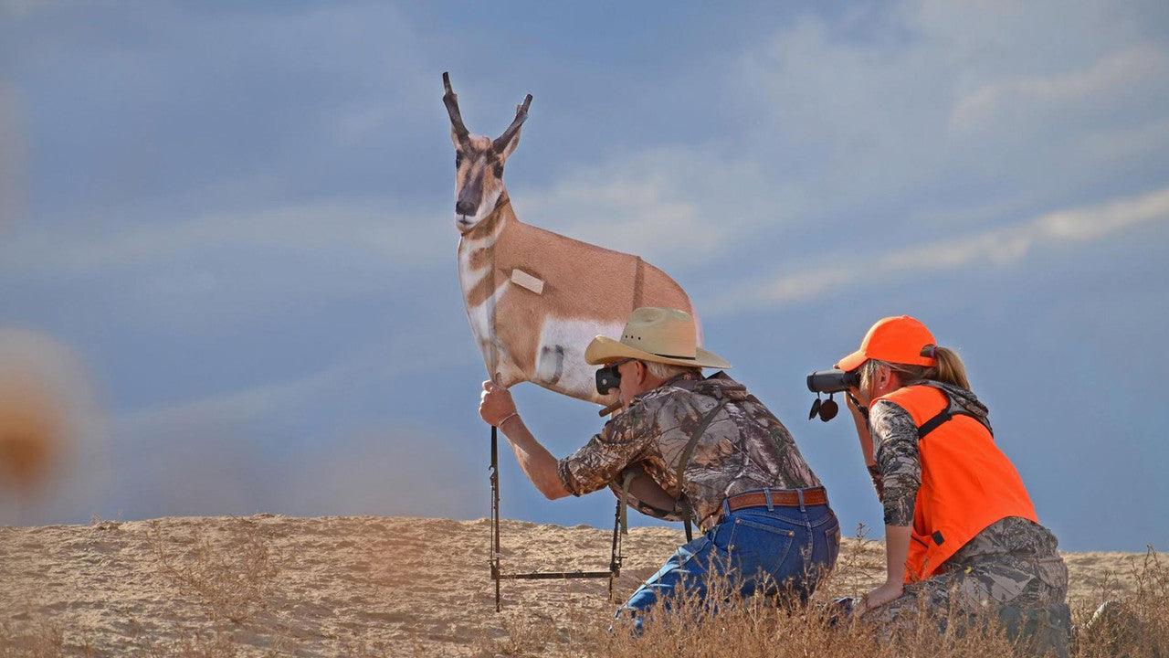 Hunters in camouflage with antelope decoy and binoculars on open plain under blue sky