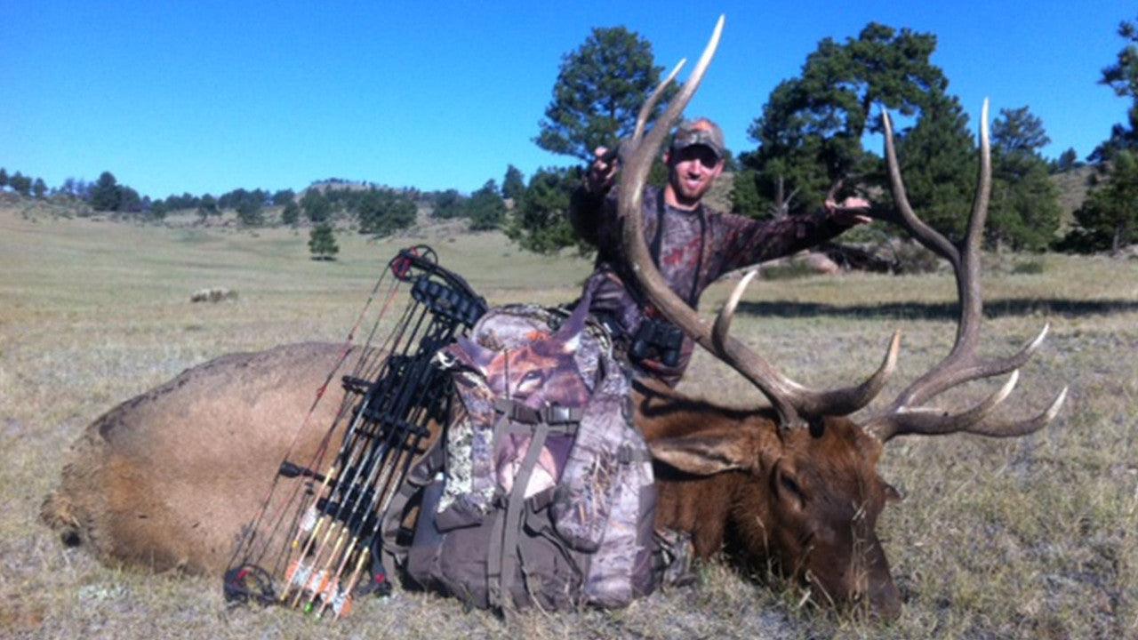 Hunter kneeling beside elk decoy with bow gear in open grassy field