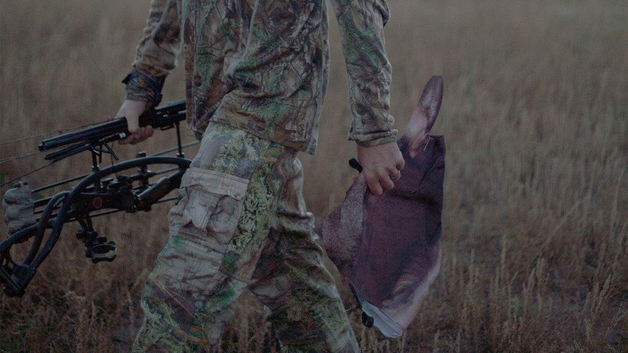 Hunter in camo carrying a compound bow and elk decoy in grassy field