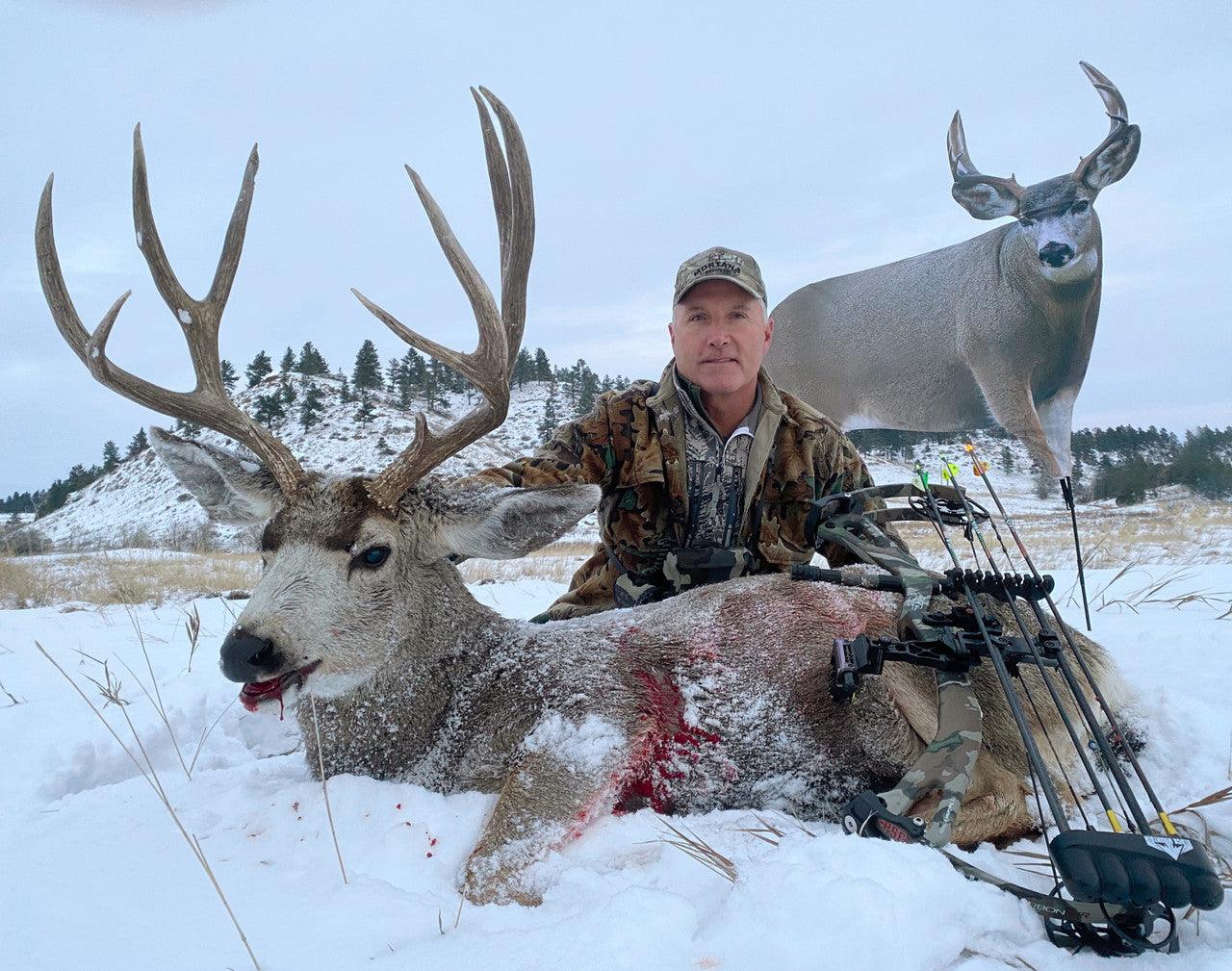 Hunter with harvested mule deer buck in snowy field, bow and arrows visible, winter hunting scene