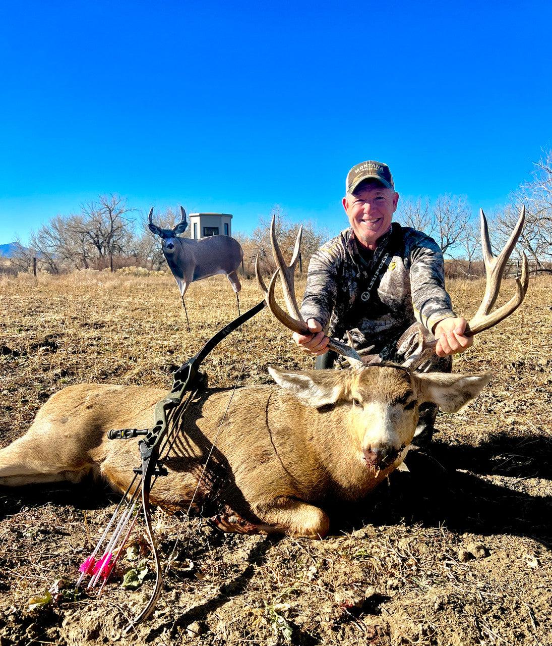 Hunter posing with harvested mule deer buck, compound bow, and decoy in open field