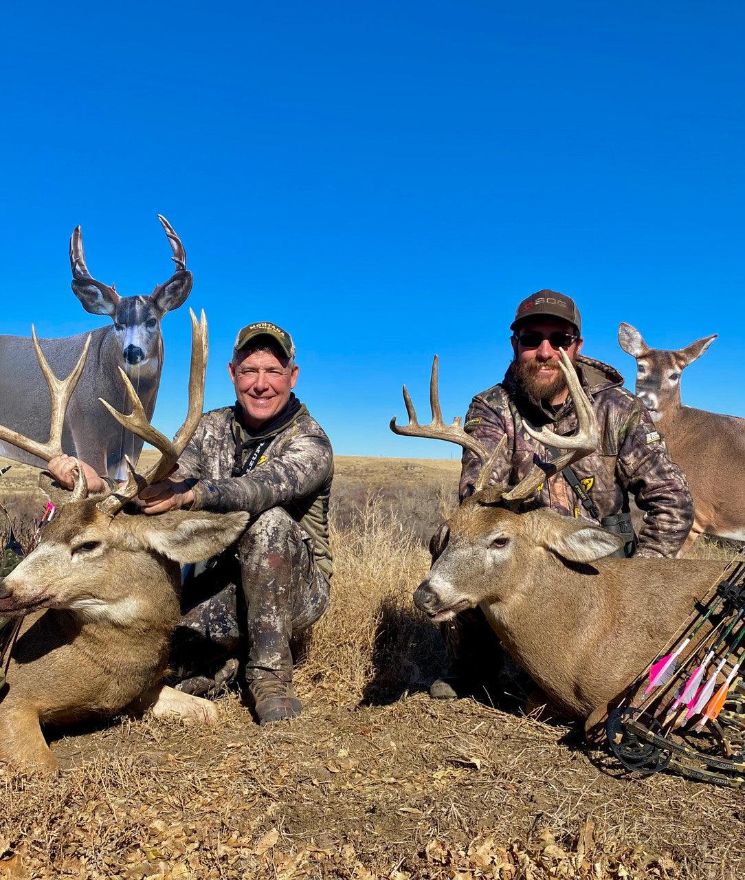 Hunters in camouflage gear kneeling with two harvested deer and archery bows in grassy field