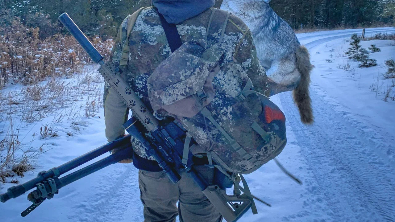 Hunter in camouflage gear carrying a rifle and a coyote on a snowy forest trail