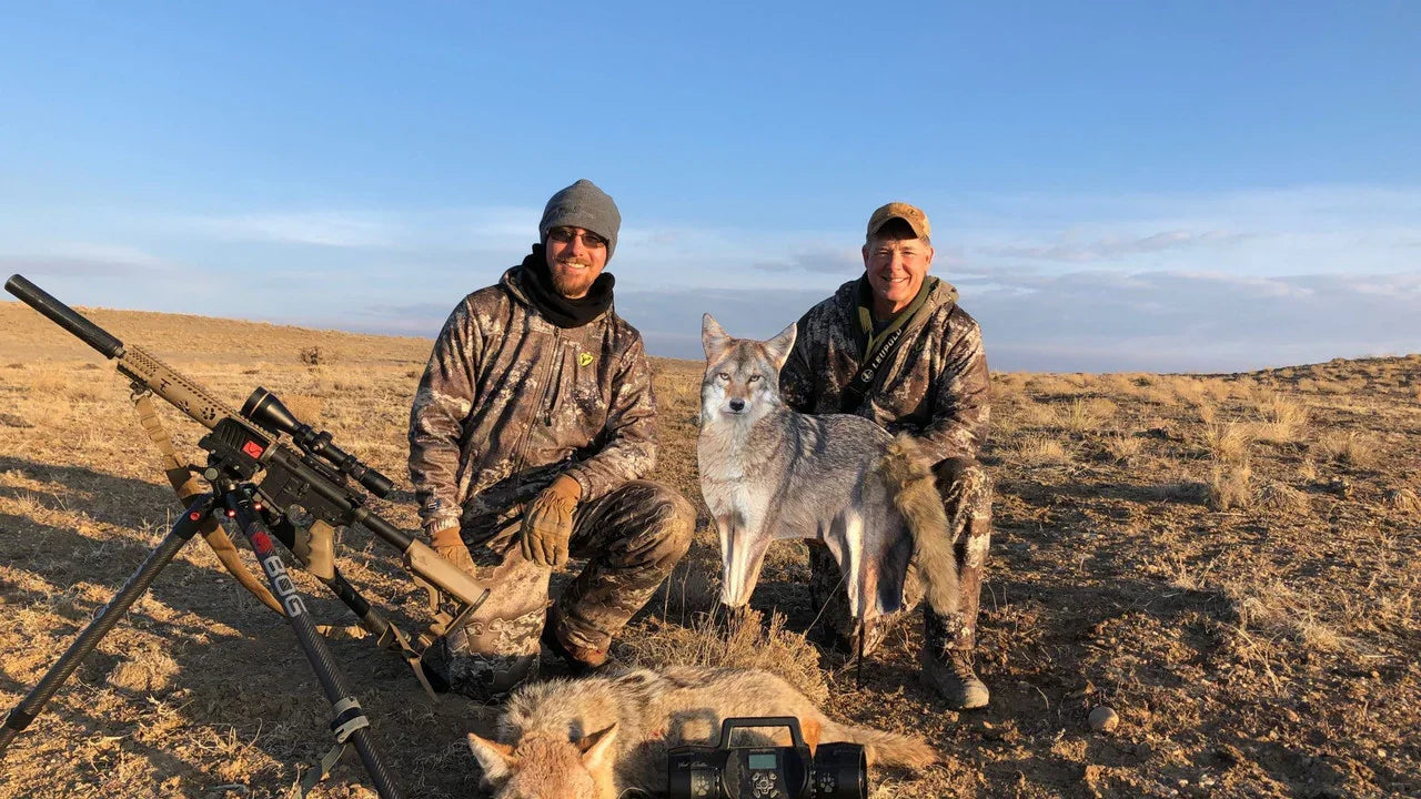 Two hunters in camo with rifles and coyote decoy on dry grassland, hunting trip scene
