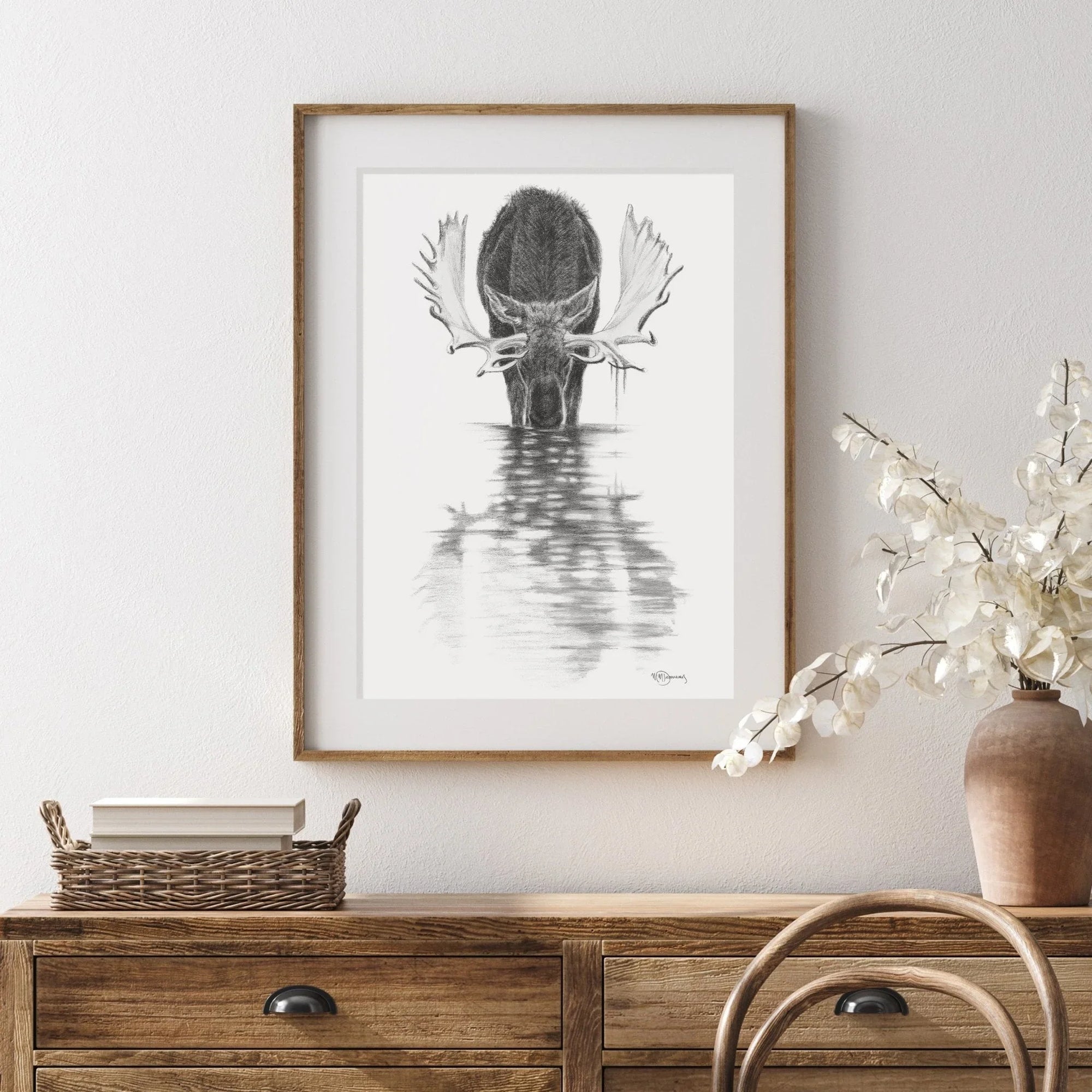 Framed black and white drawing of moose in calm water, displayed above wooden dresser