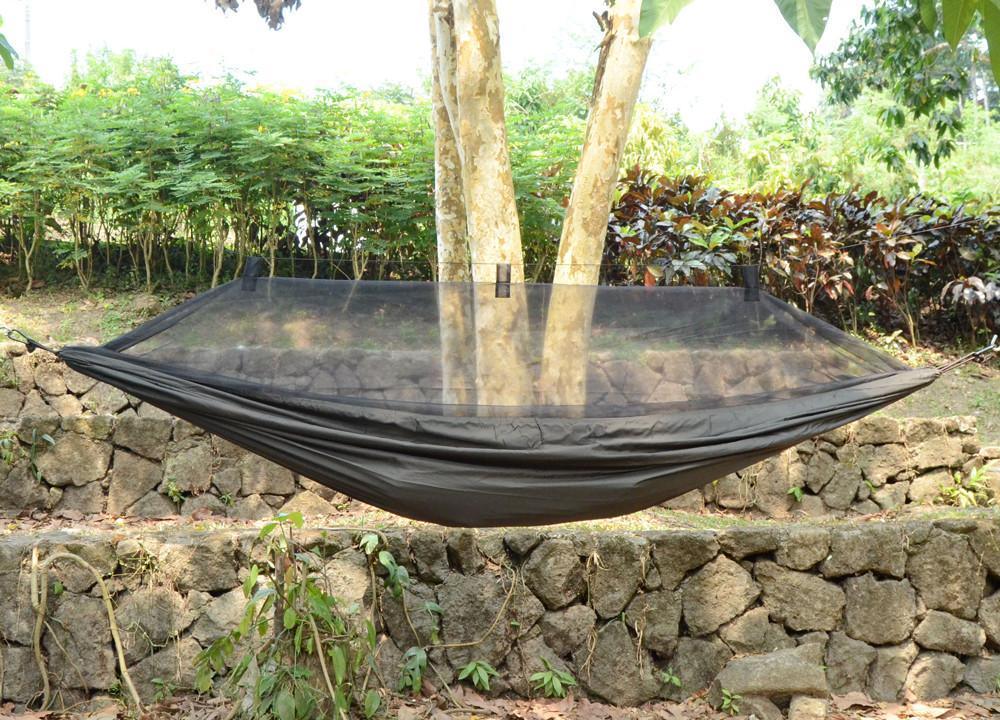Black camping hammock with mosquito net hung between trees outdoors near stone wall