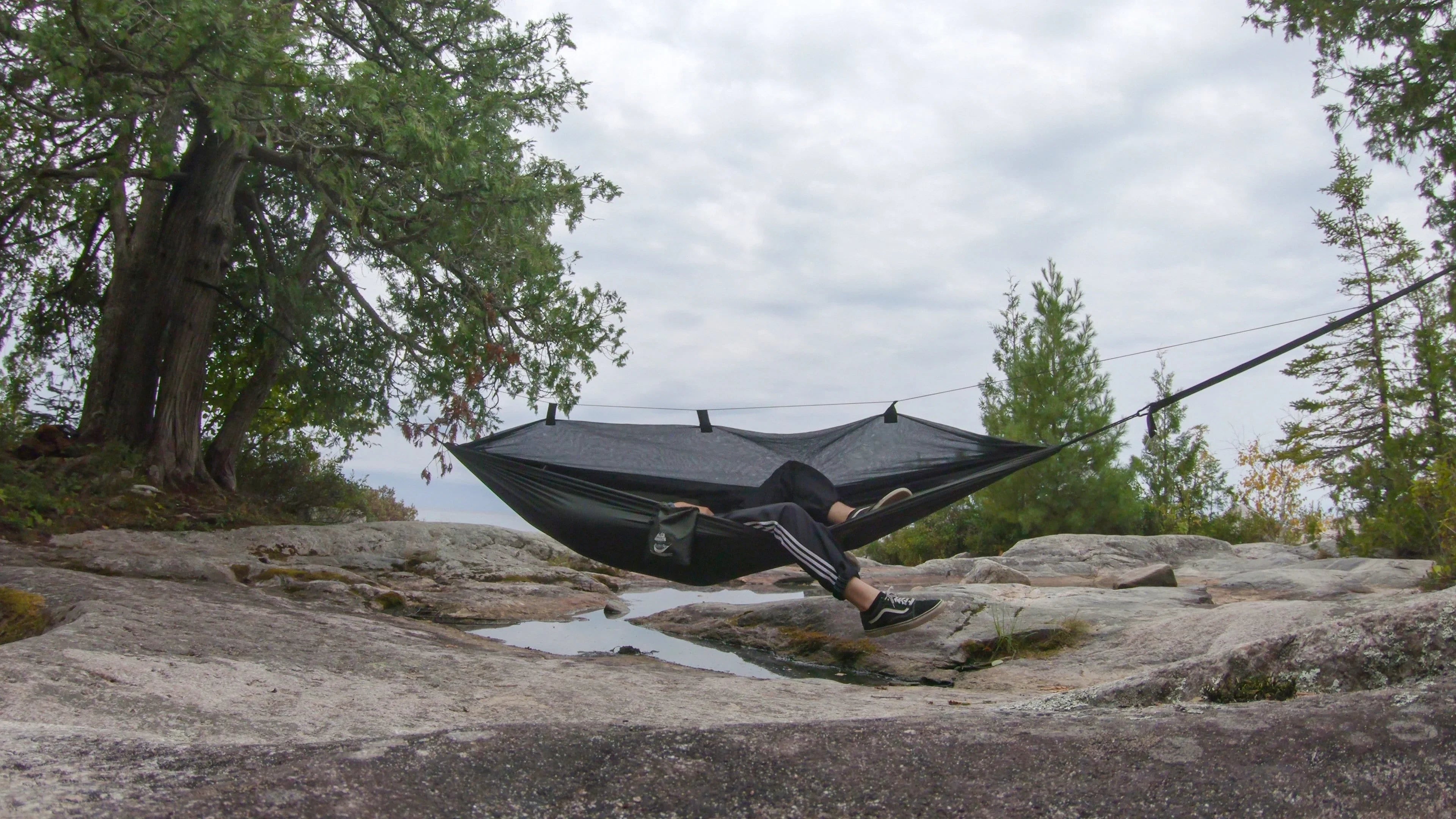 Person relaxing in a black mosquito hammock outdoors between trees on rocky terrain