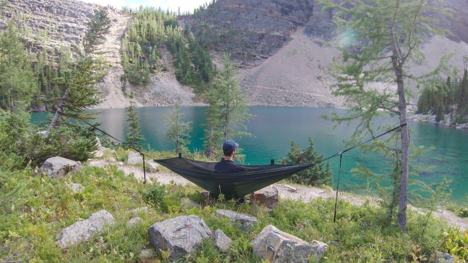 Person relaxing in a hammock by a forest lake with mountains and pine trees