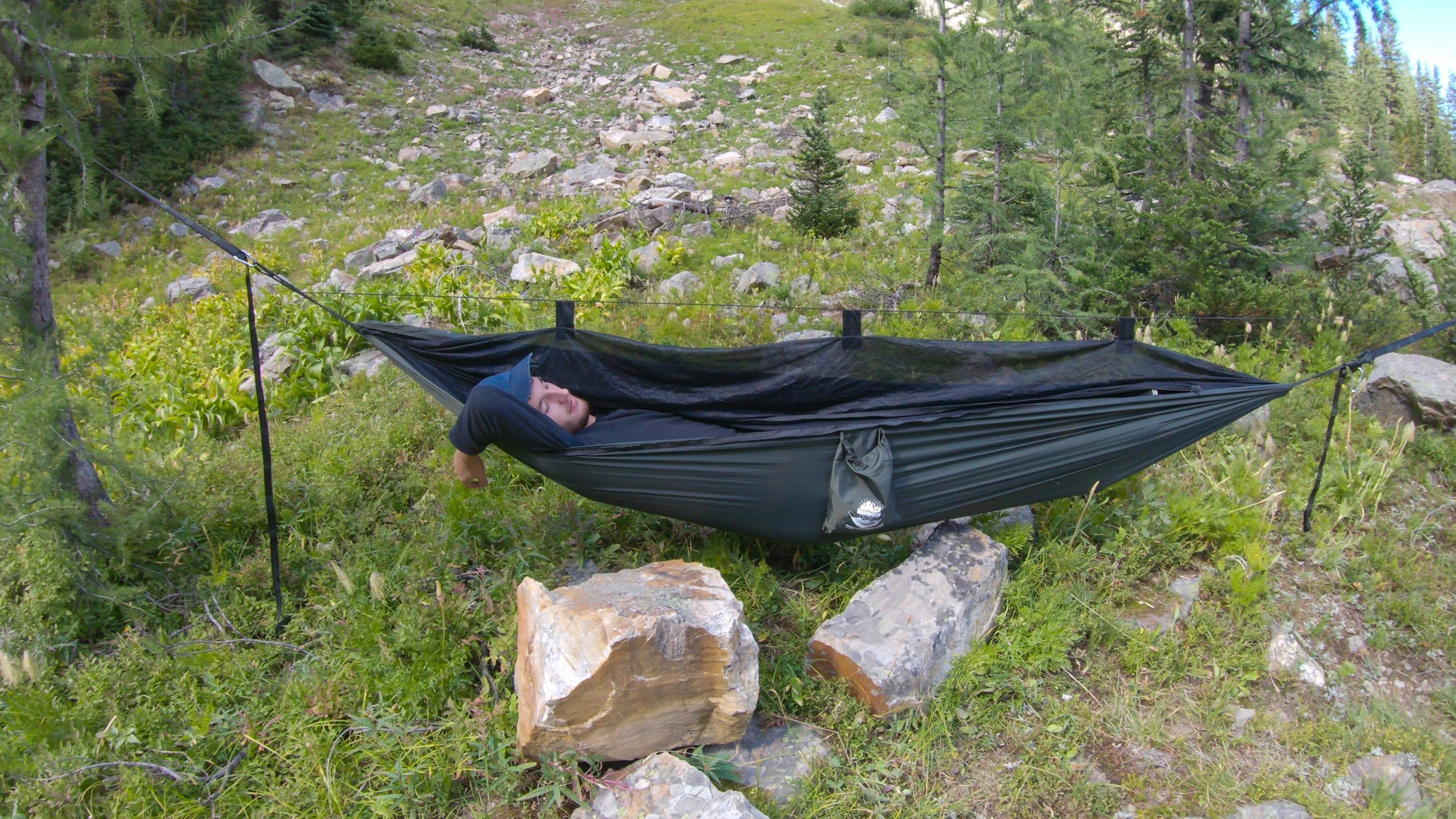 Man relaxing in a black mosquito hammock with bug net in a rocky forest campsite