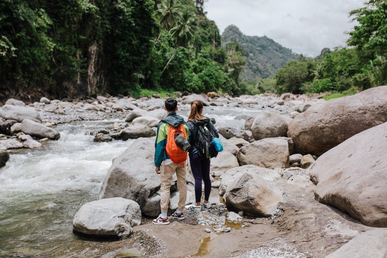 Two hikers with backpacks standing on rocks by a river in a lush green forest