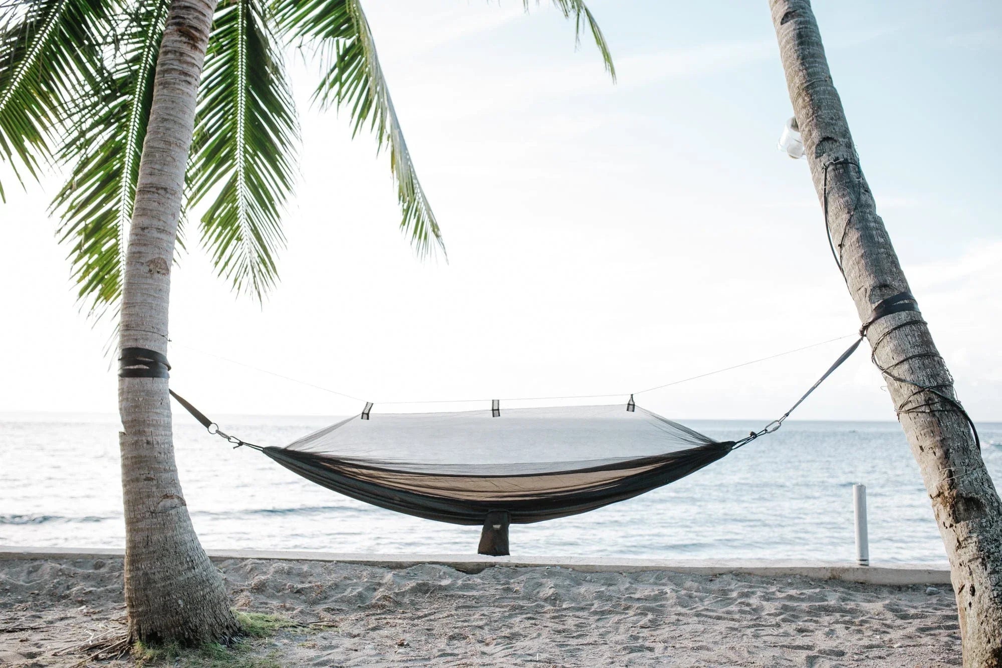 Beach hammock with mosquito net between palm trees by the ocean on sandy shore