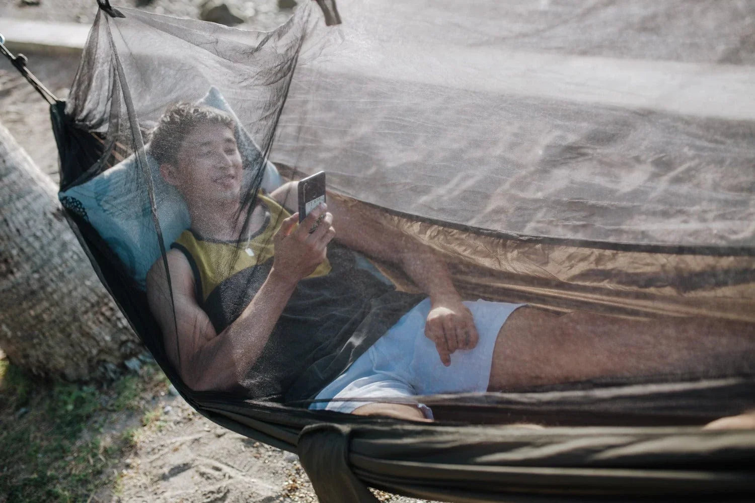 Man relaxing in a mesh hammock outdoors using smartphone near trees